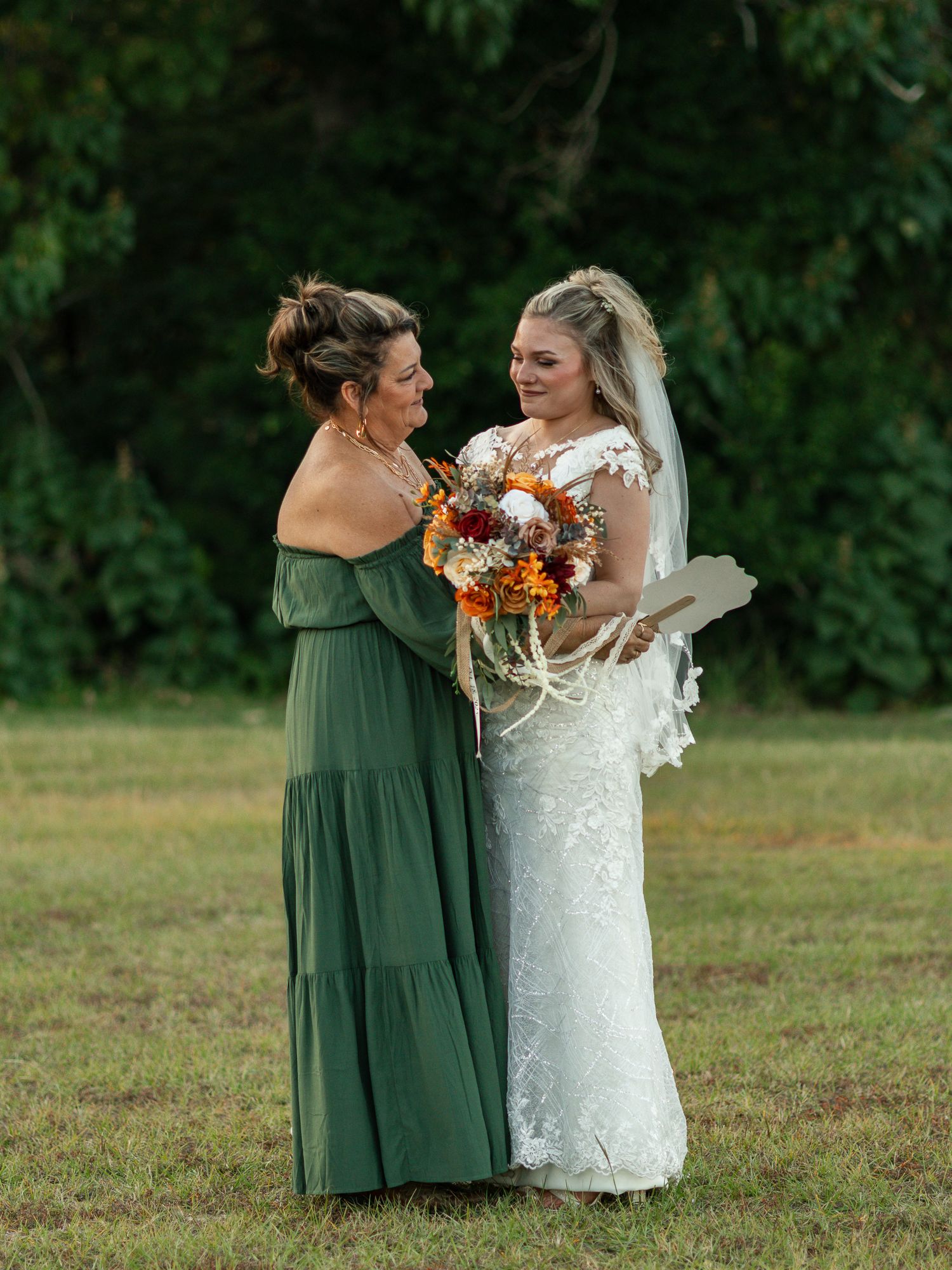 A bride in a lace gown and bridesmaid in an olive green dress share a moment while holding a fall-colored bouquet.