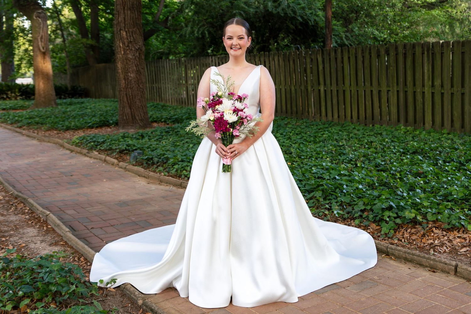 Bride in classic white ballgown with train holding pink and burgundy bouquet on brick pathway.