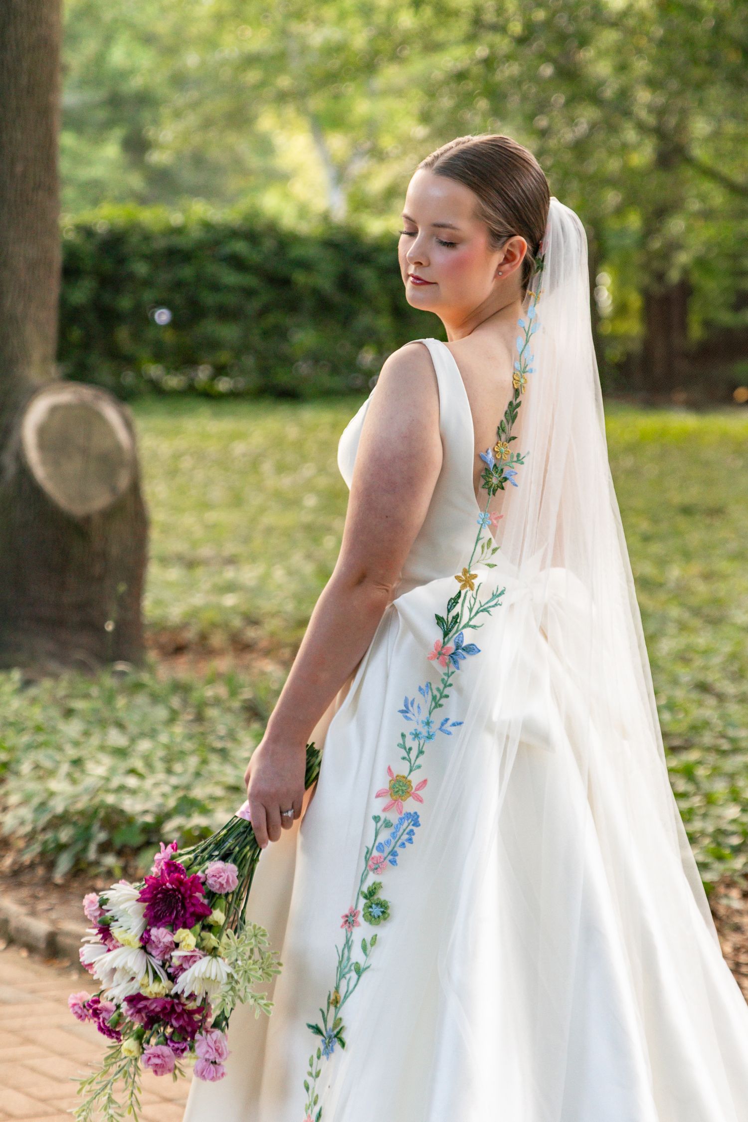 Bride in white gown with floral embroidery and veil holding a purple and white bouquet outdoors.