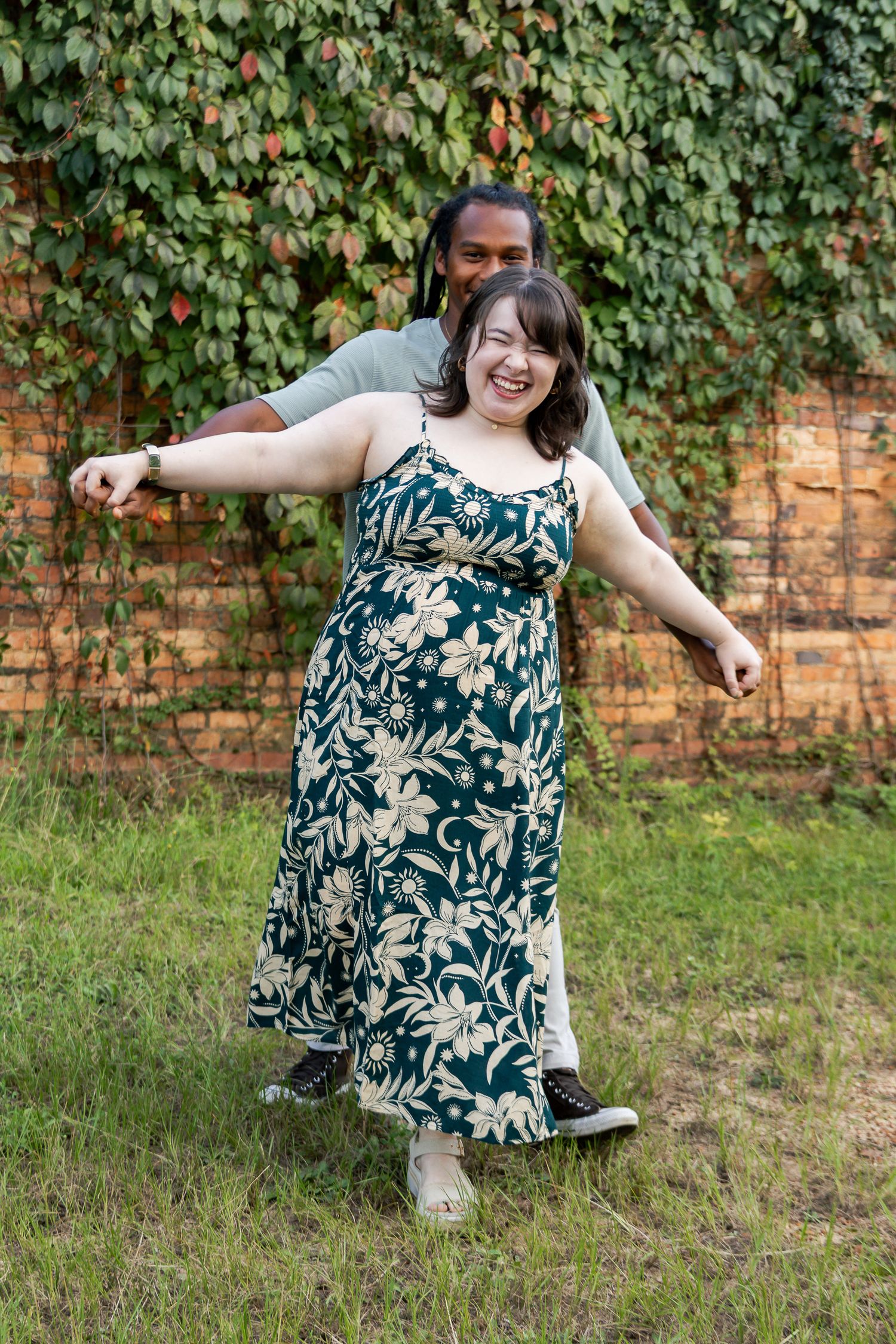 Joyful woman in floral dress being spun by her partner in front of an ivy-covered brick wall.