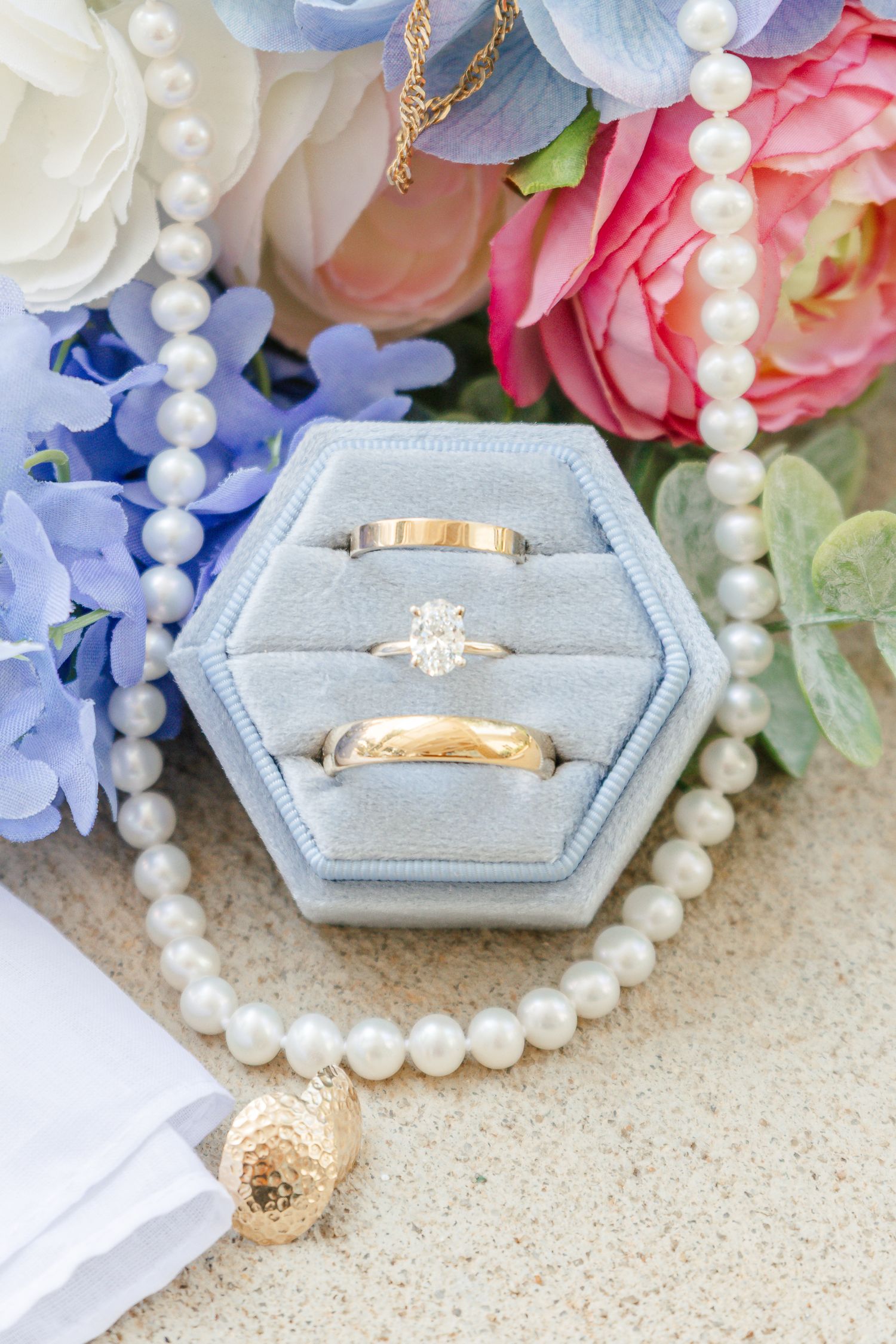 Gold wedding rings and diamond engagement ring in blue velvet hexagon box with pearl necklace and floral bouquet.