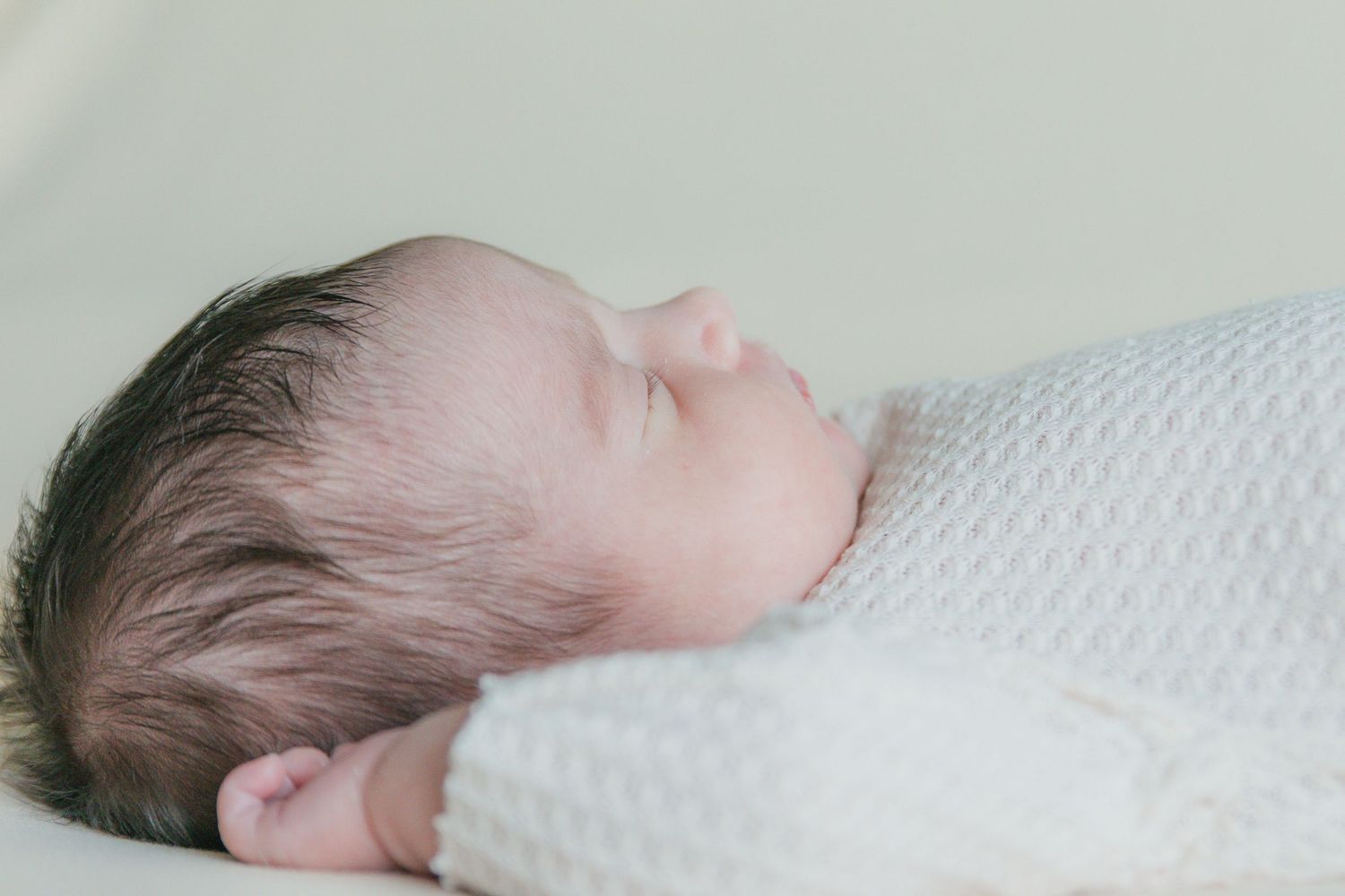 Sleeping newborn baby swaddled in white knit blanket with dark hair, peaceful side profile view.