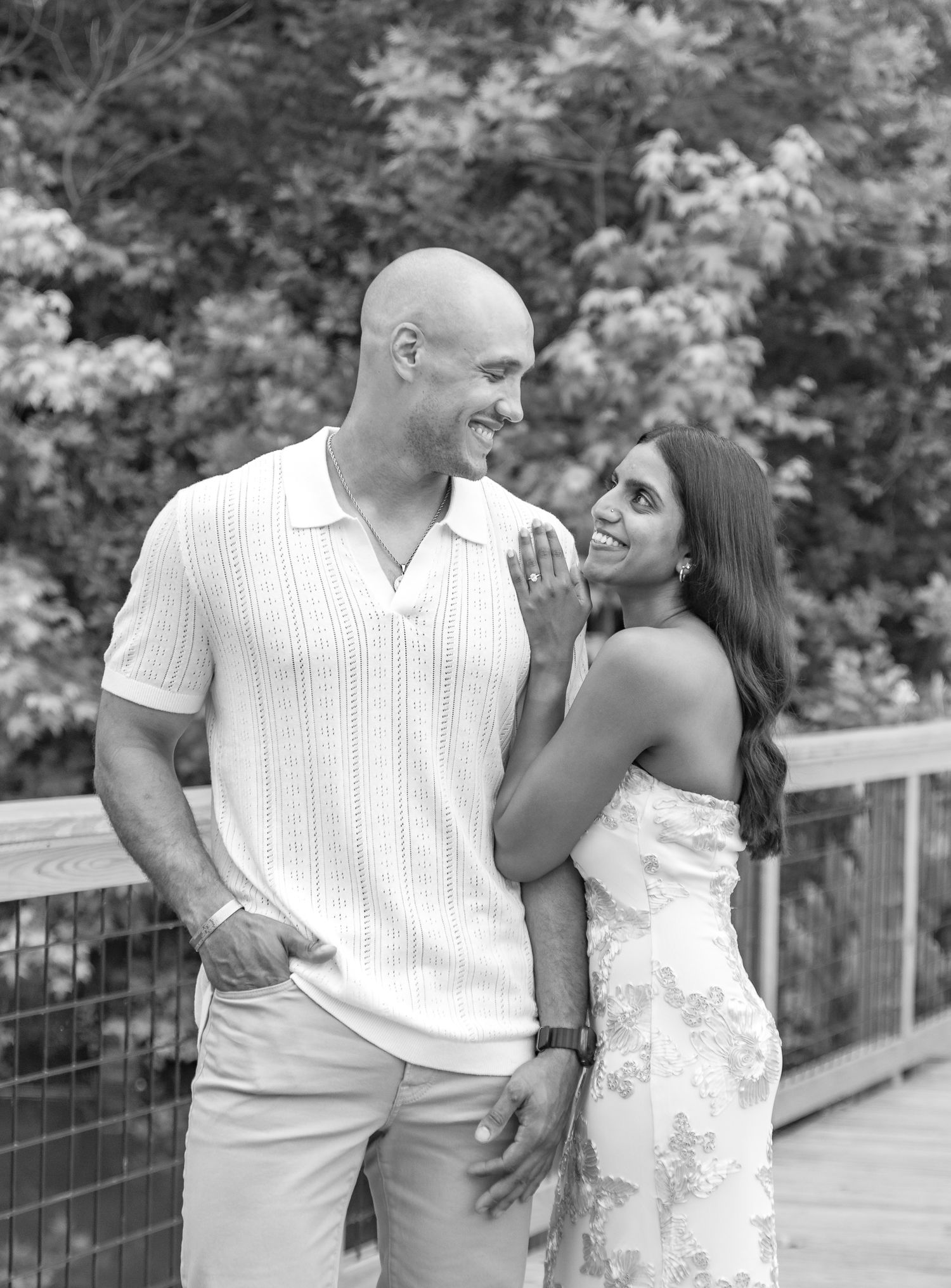 Black and white portrait of a couple smiling at each other on an outdoor wooden bridge.