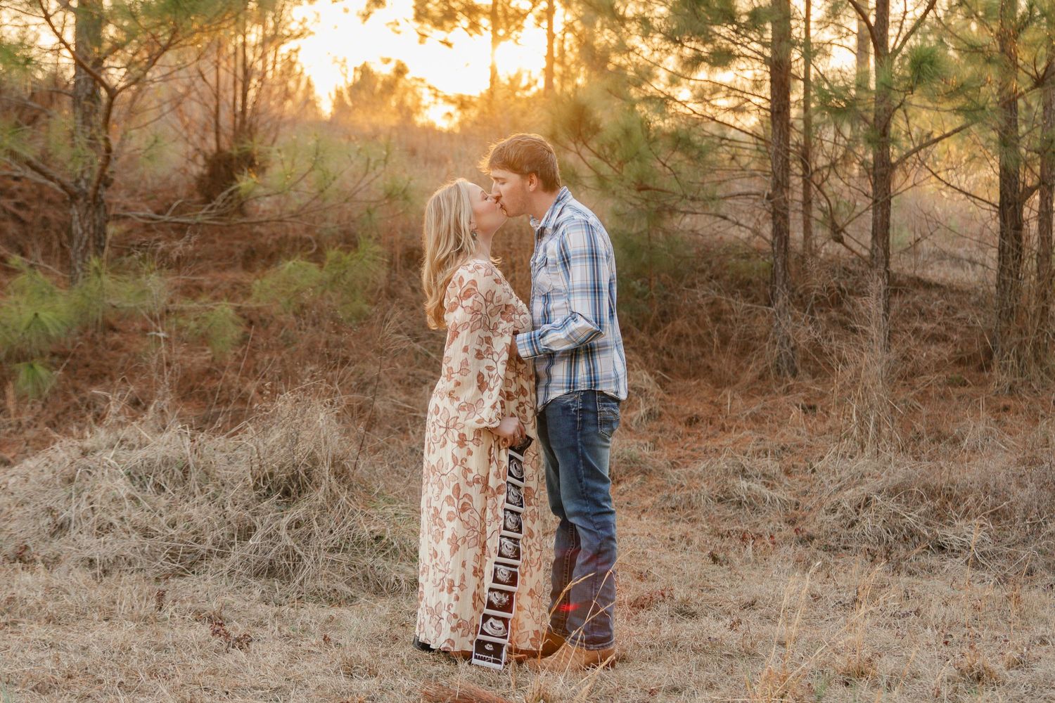 A couple in western attire shares an intimate moment at sunset in a pine forest setting.