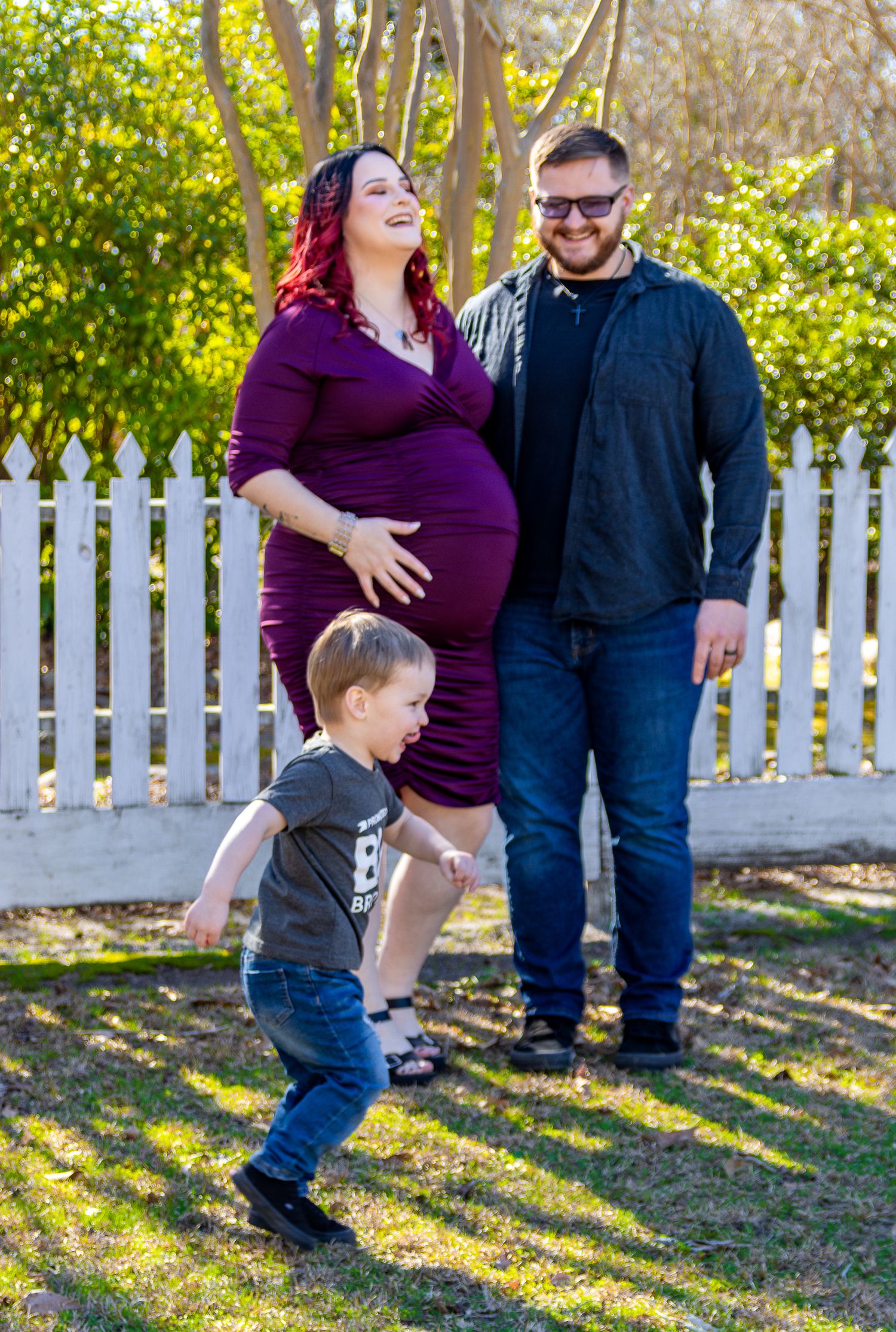 A pregnant woman in a purple dress stands with her partner and young son near a white picket fence outdoors.
