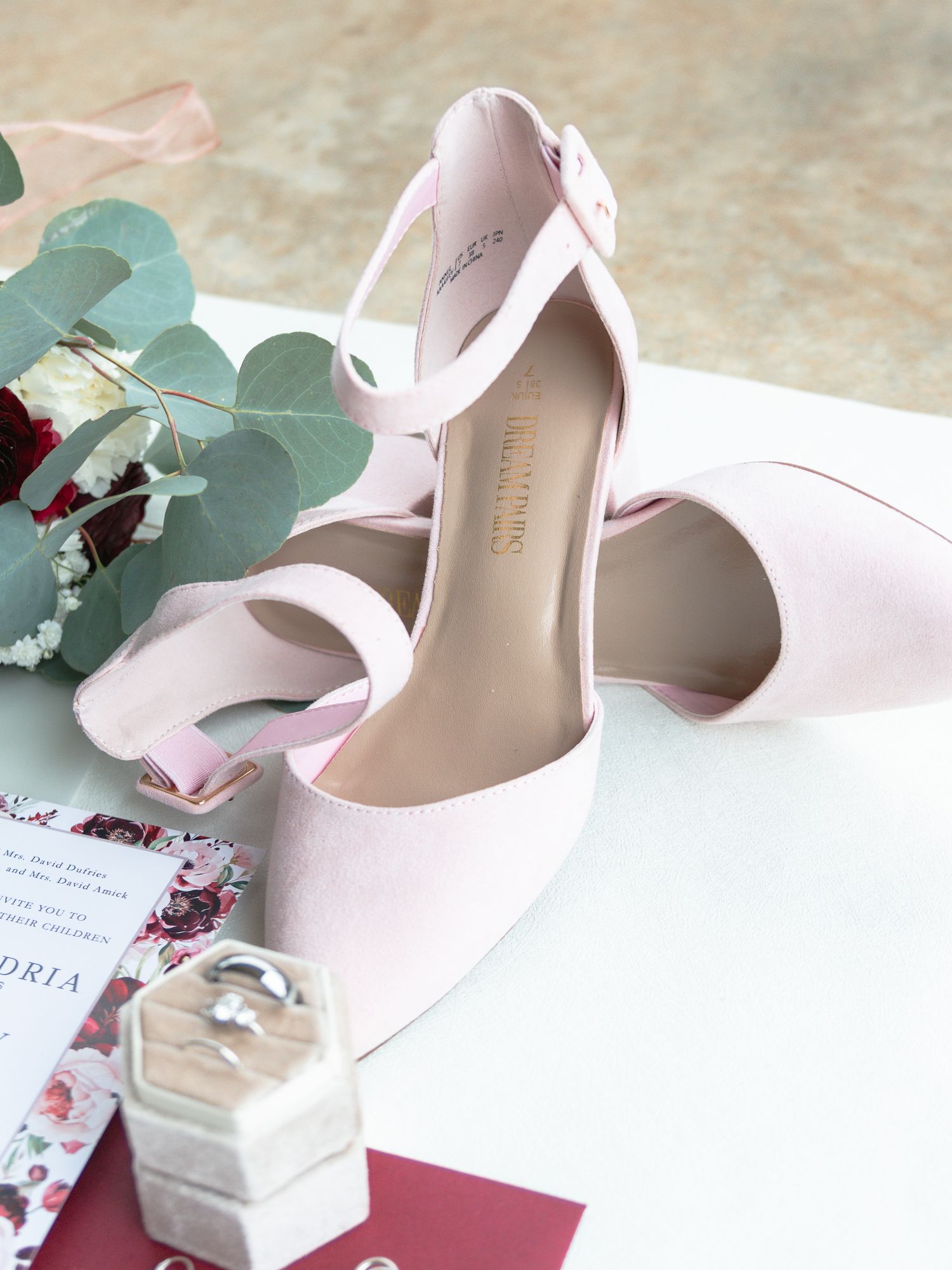Elegant nude high heel wedding shoes with eucalyptus leaves and floral decorations arranged on white surface.