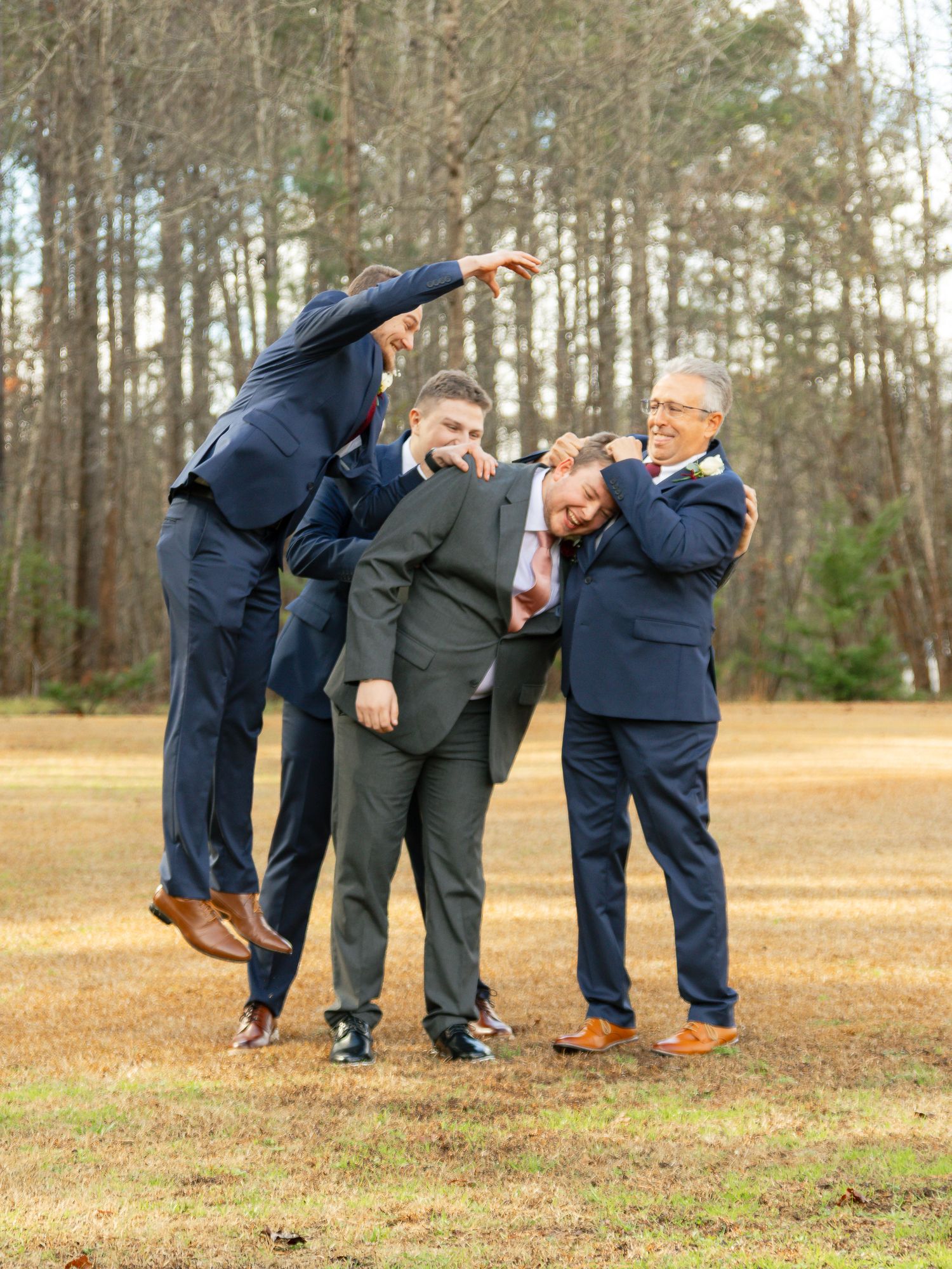 Four people in navy and gray suits pose playfully outdoors in a wooded area with brown grass.