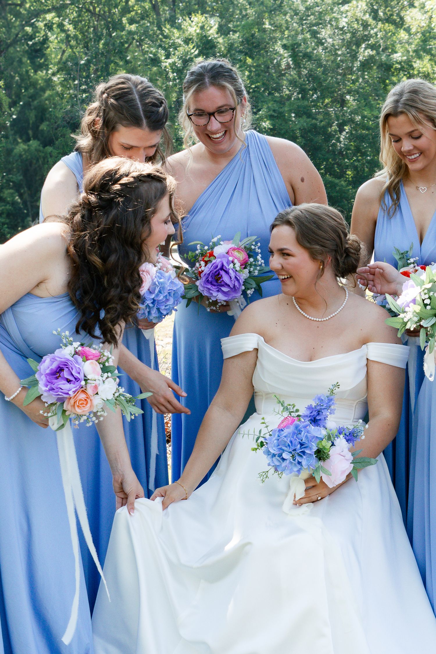 Bridesmaids in light blue dresses and bride in white gown share joyful moments while holding colorful floral bouquets.