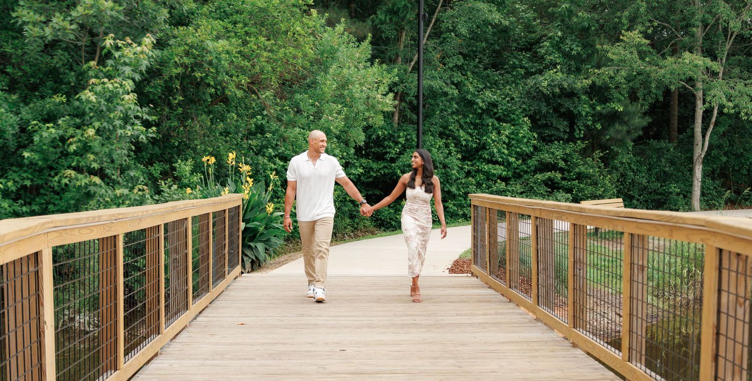 Couple holding hands walking across a wooden bridge surrounded by lush green trees.