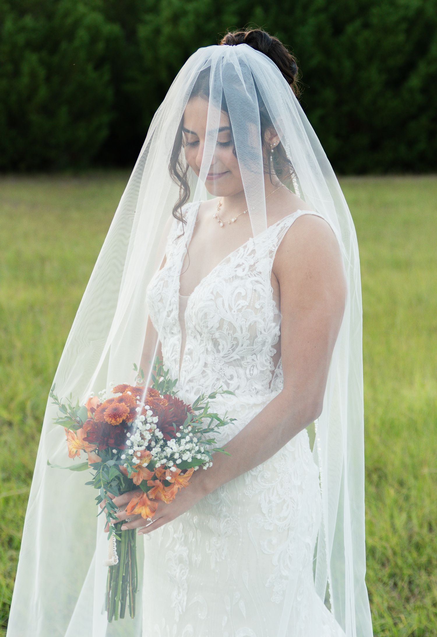 A bride in a lace wedding dress with a flowing veil holds a colorful bouquet while standing in a sunlit meadow.