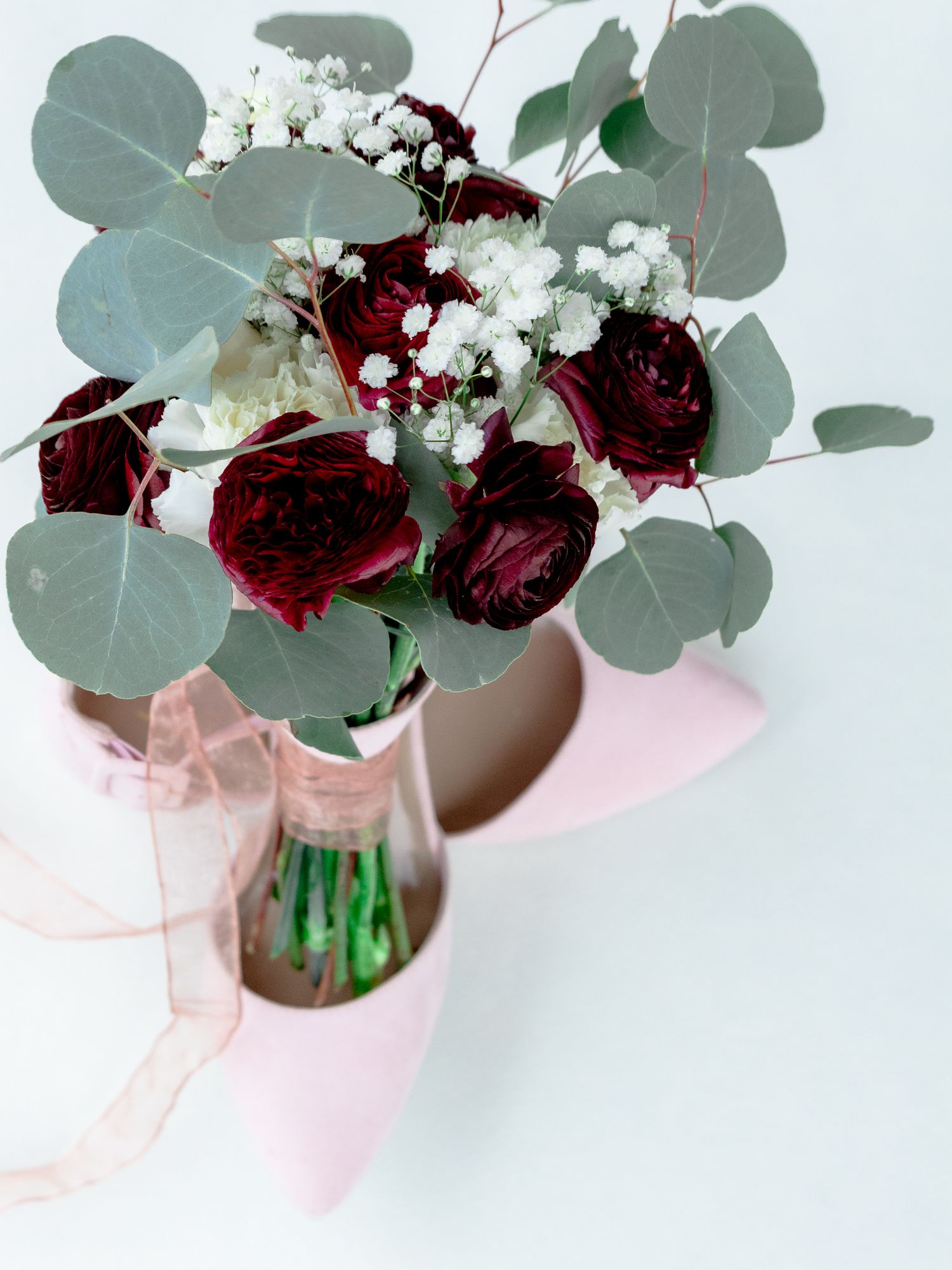 Wedding bouquet with deep burgundy roses and baby's breath tied with pink ribbon against light background.