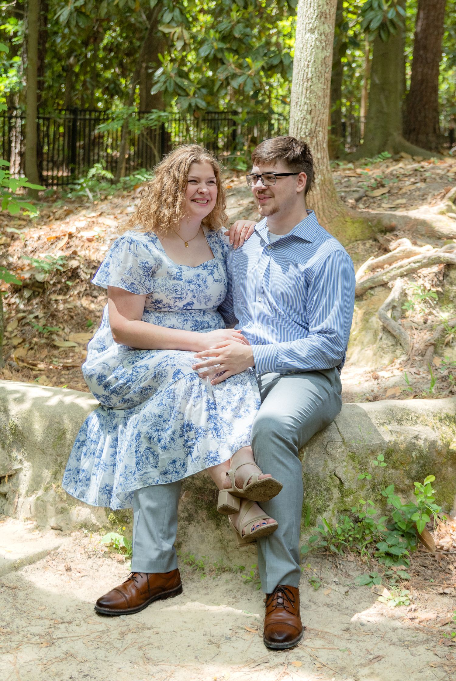 A smiling couple sits together on stone steps in a wooded park, dressed in coordinating blue outfits.