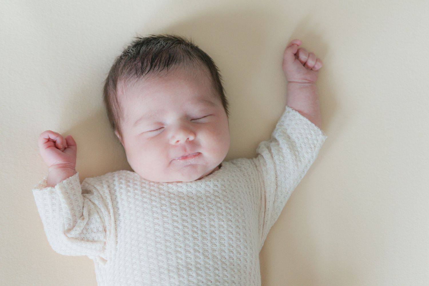 Peaceful newborn baby sleeping with arms raised in a white knit onesie on a soft cream background.