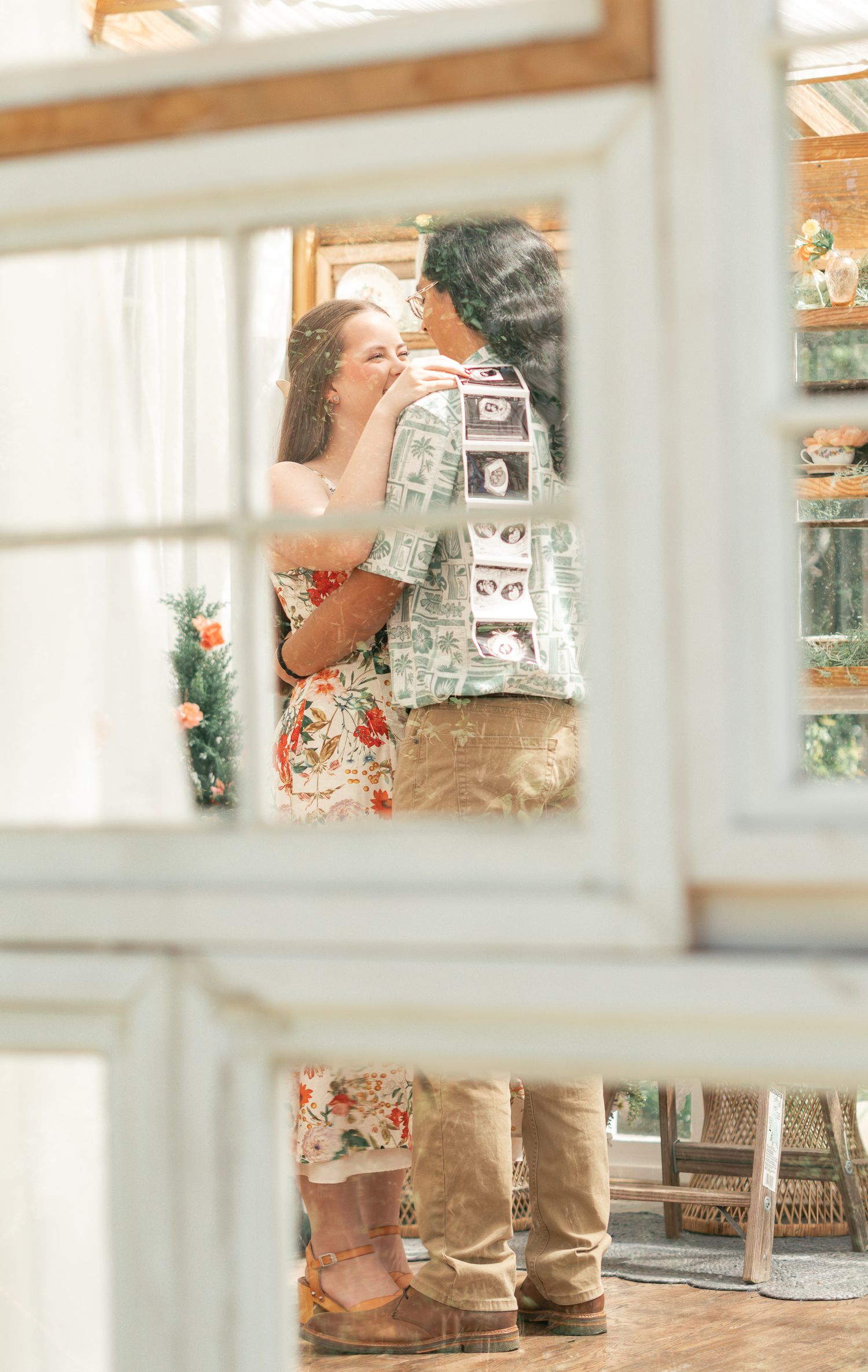 Couple sharing a tender moment viewed through white window frames, with ultrasound photos on shirt.