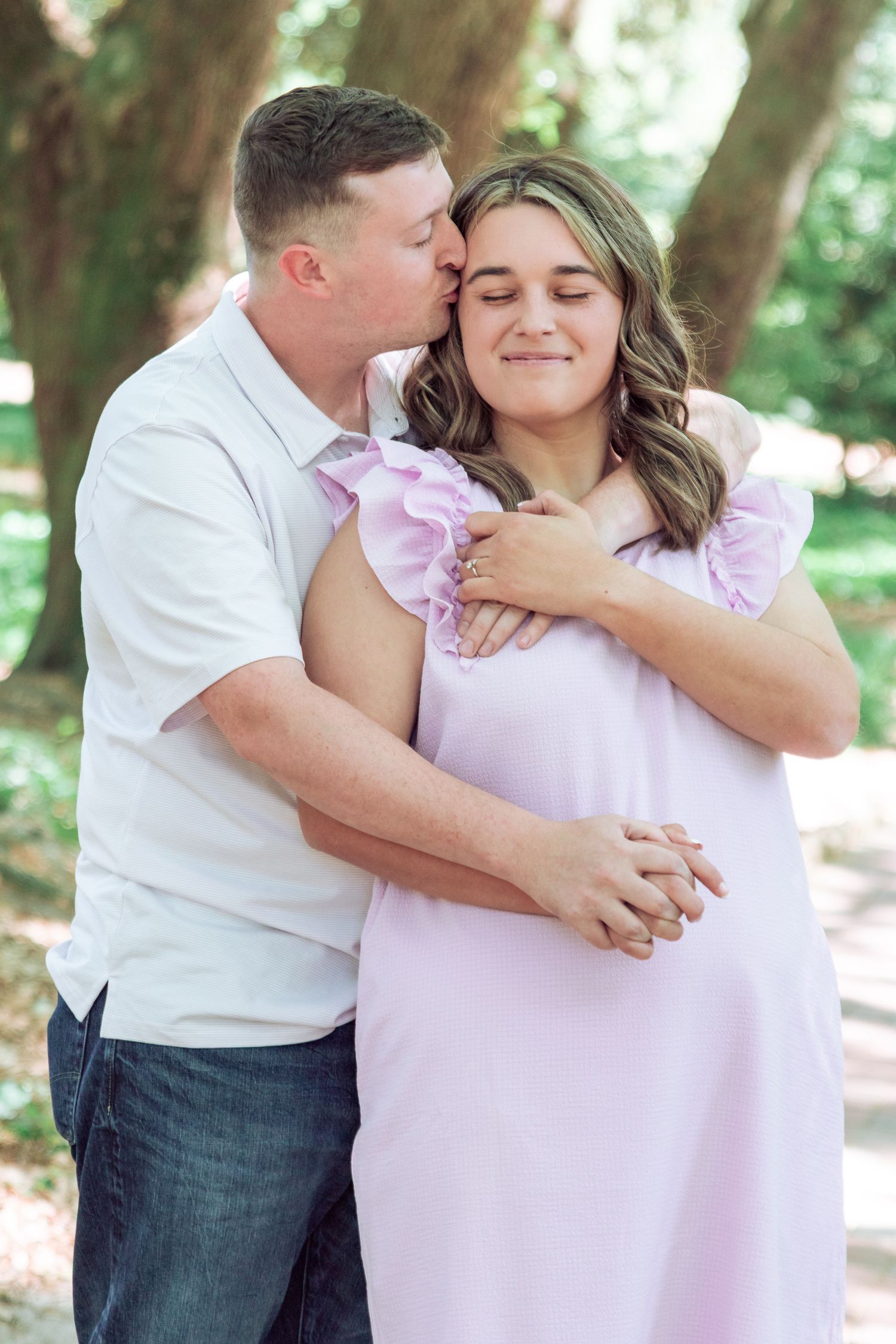 A man in a white shirt kisses a pregnant woman in a lavender dress outdoors among trees.