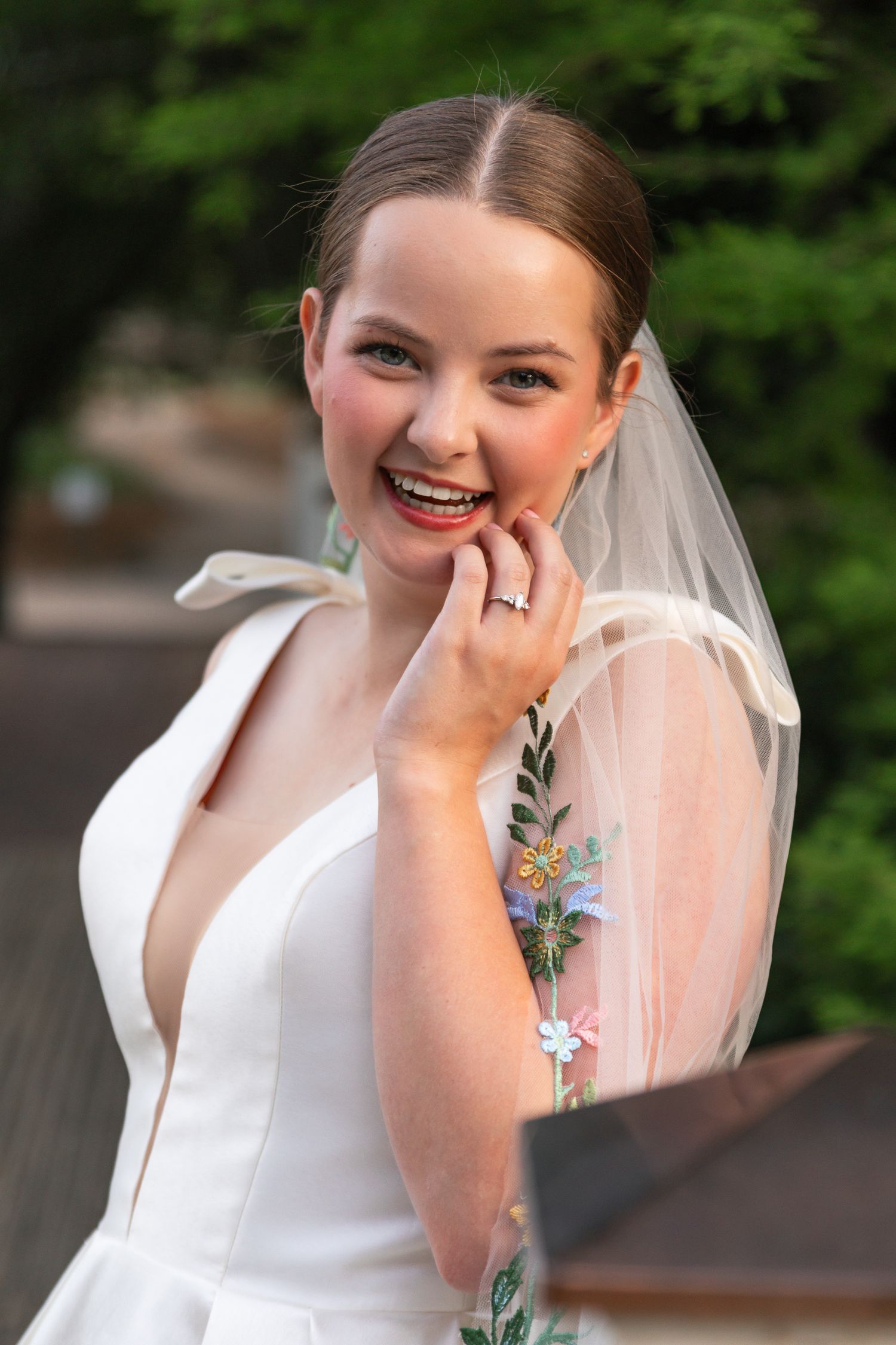 Laughing bride in deep V-neck white gown with floral embroidered veil touching her chin outdoors.