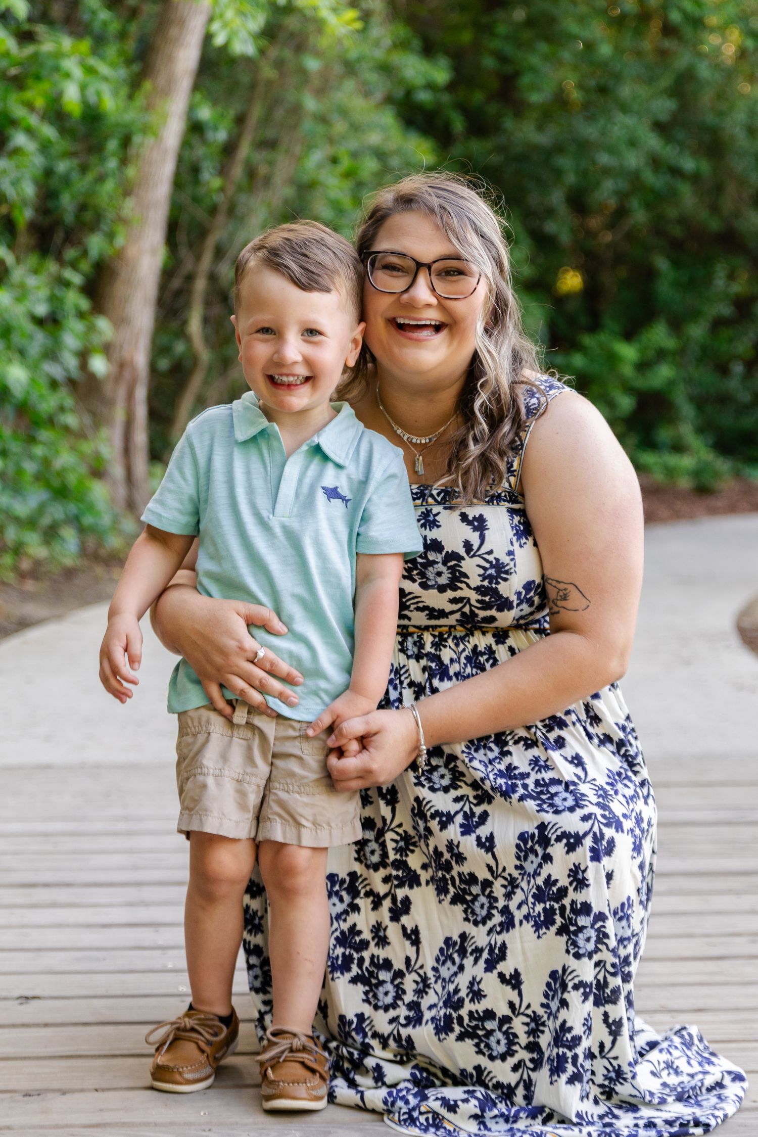 A laughing woman in a floral dress kneels beside a smiling young boy in a mint polo shirt on a wooden deck outdoors.