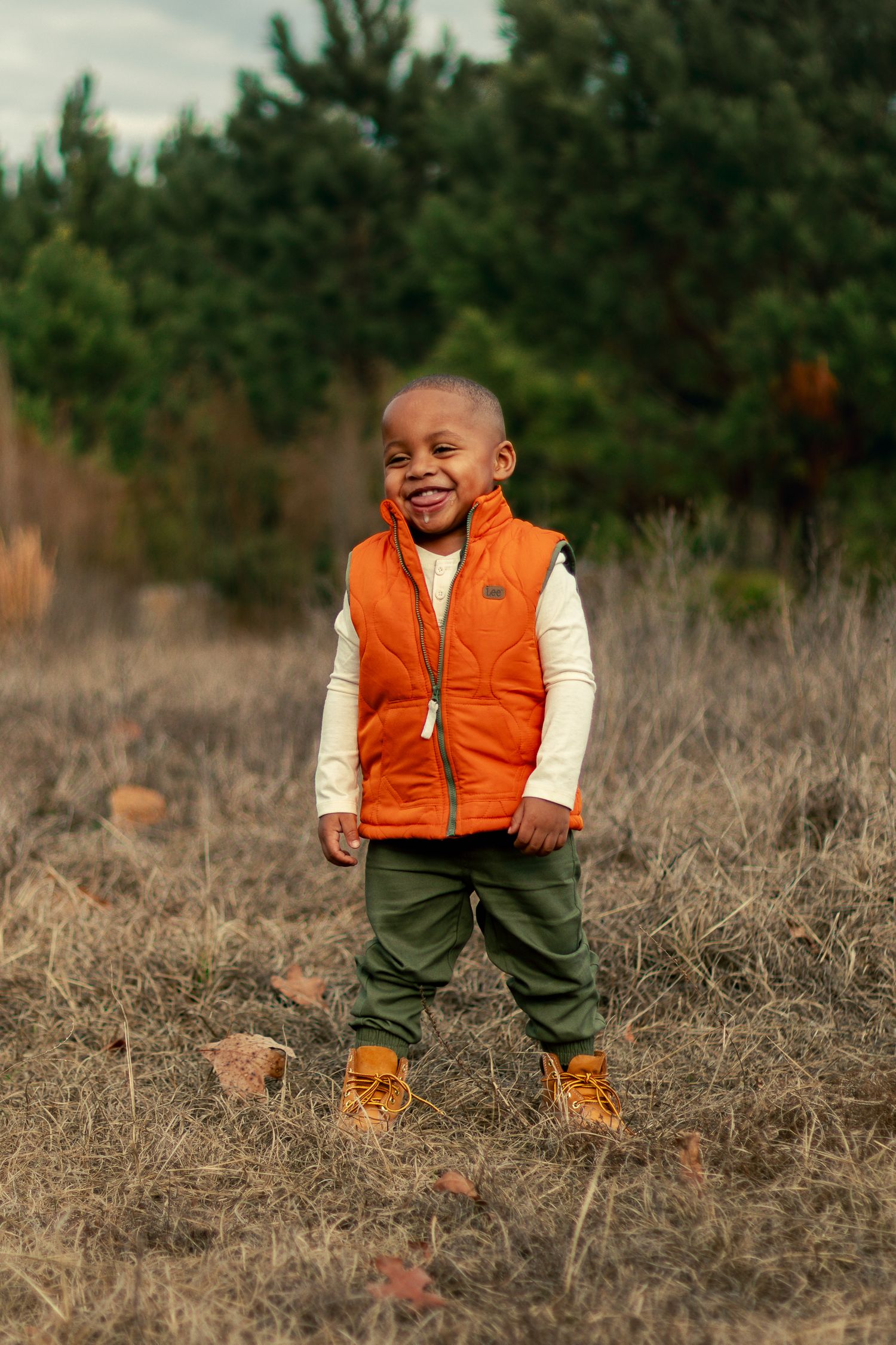 A young child in an orange vest and green pants smiles joyfully while standing in a field of dry grass.