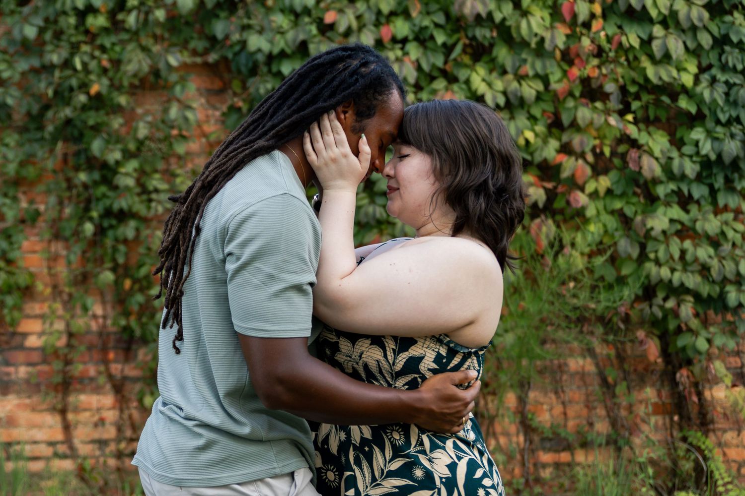 A romantic couple embraces in front of a brick wall with green ivy climbing up the background.