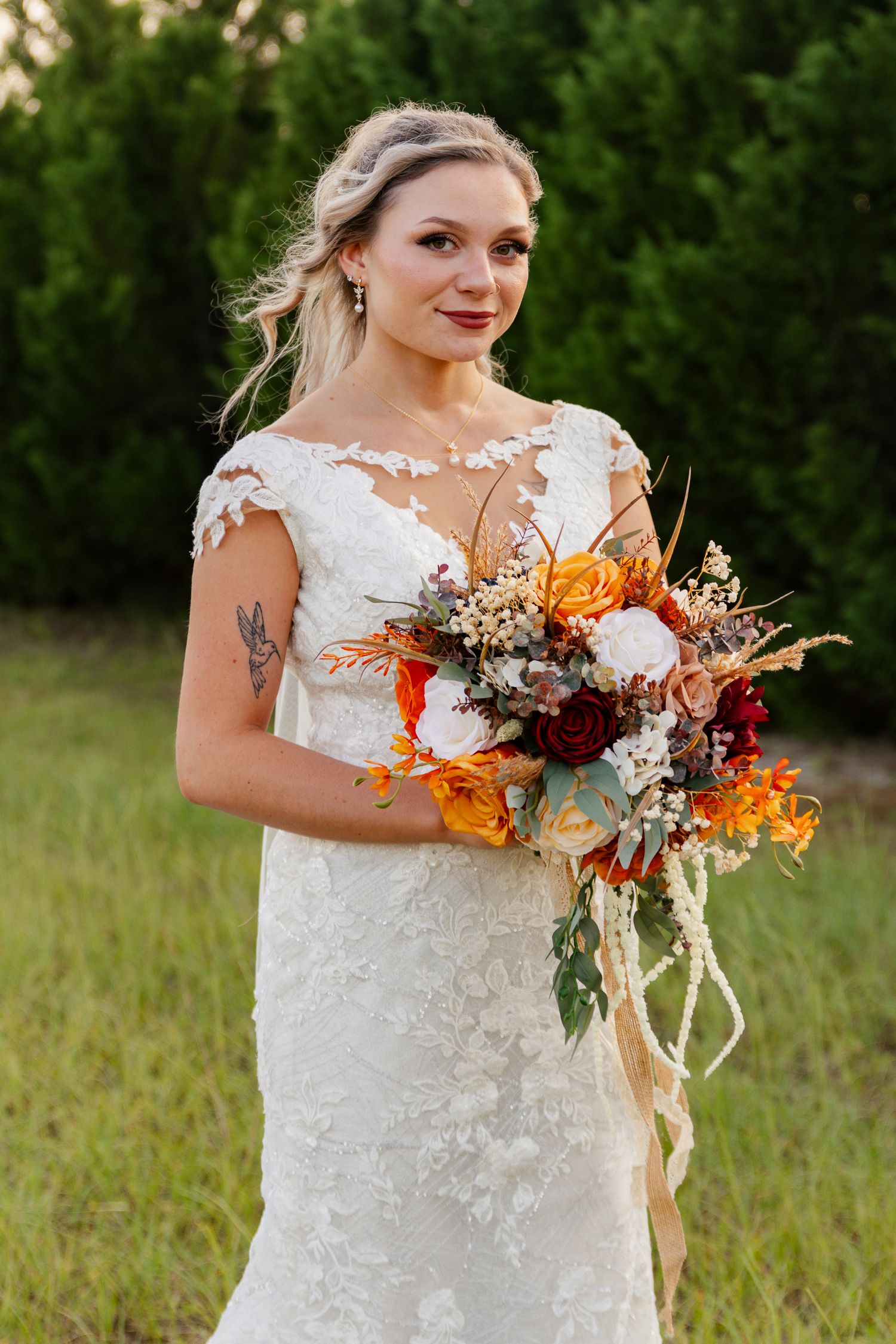 A bride in a white lace wedding dress holds a vibrant autumn bouquet with orange and burgundy flowers in a field.
