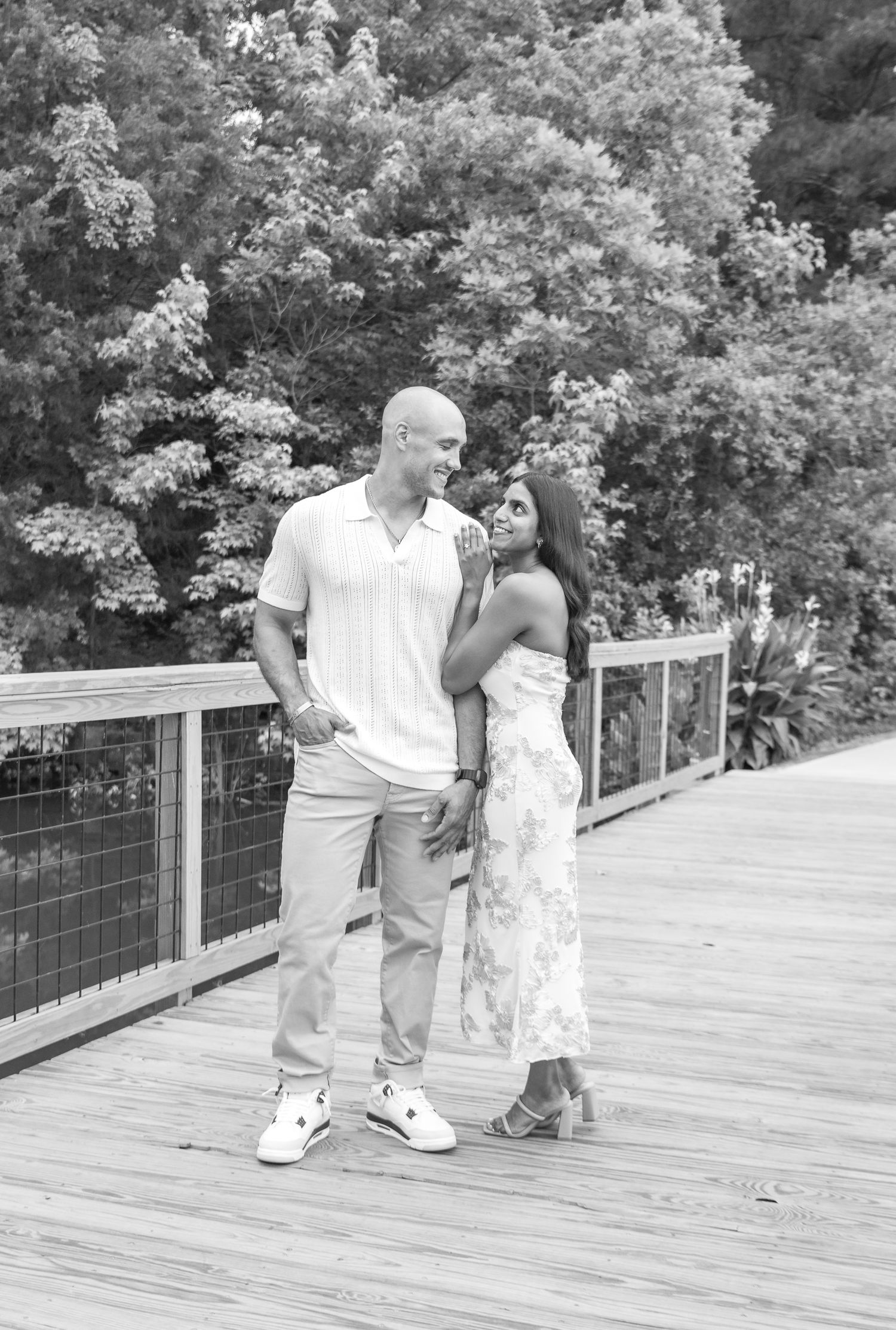 Black and white photo of a couple standing on a wooden bridge, smiling at each other with lush trees in the background.