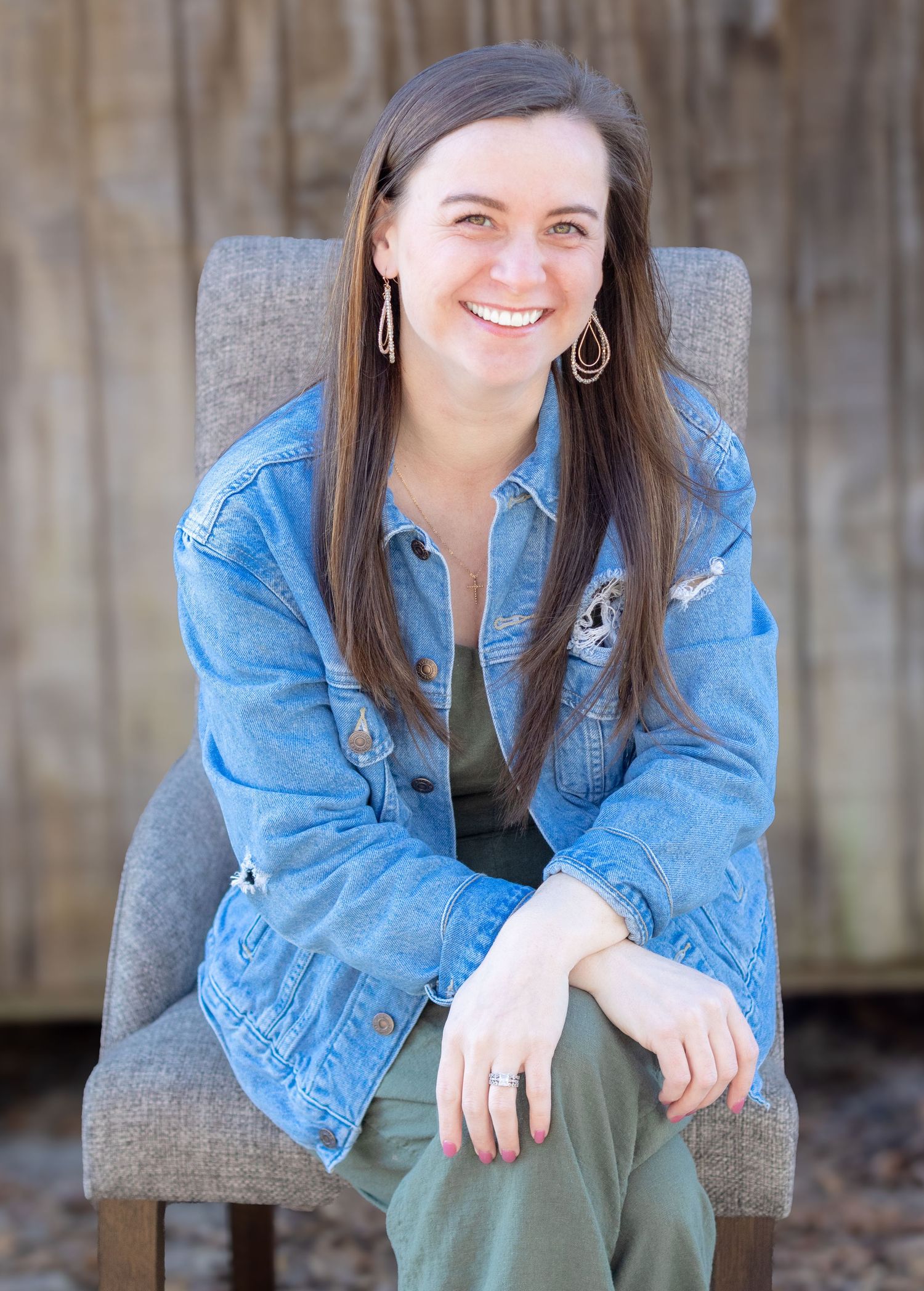 Smiling brunette woman in denim jacket seated in a gray chair against a wooden backdrop.