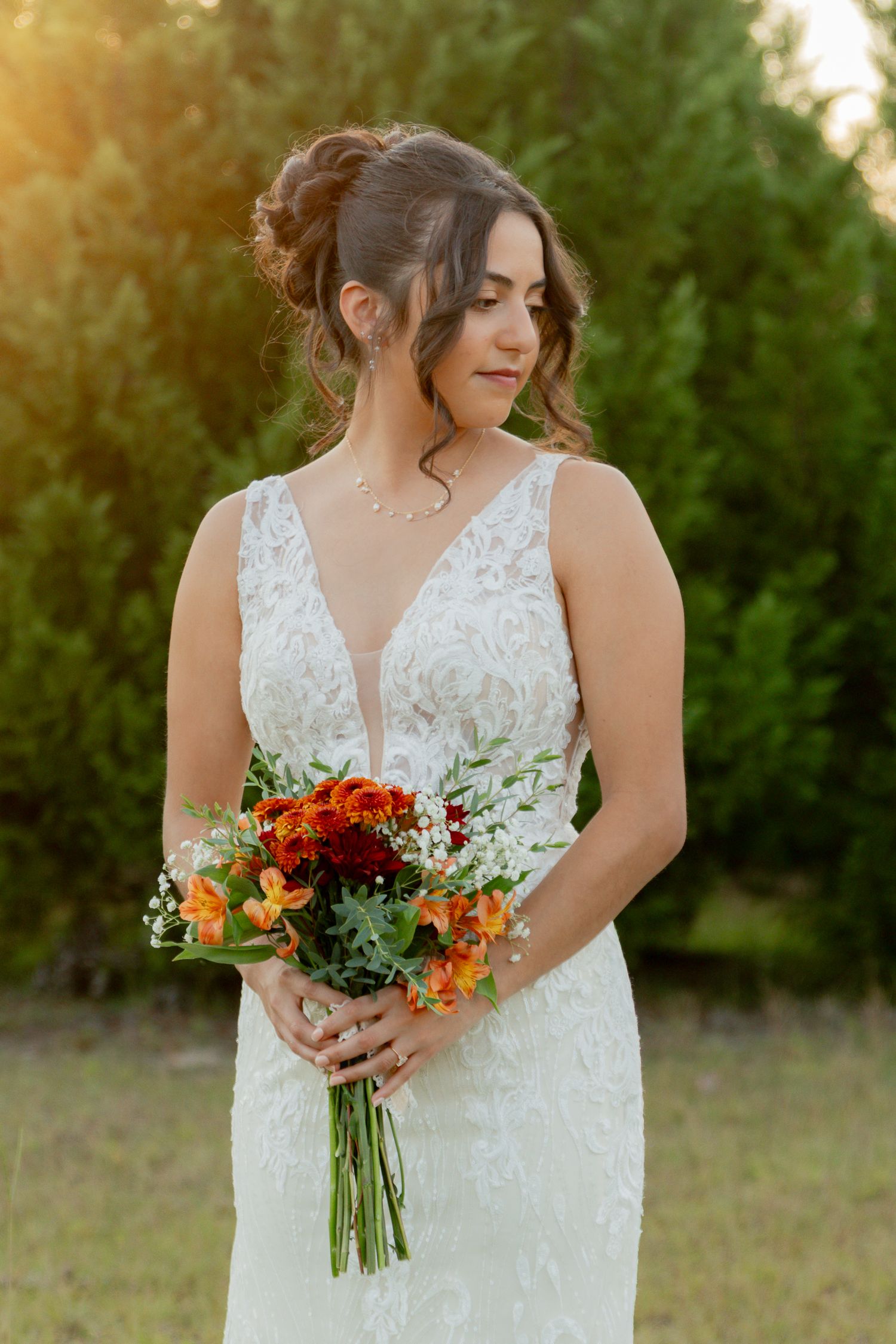 A brunette bride in a lace V-neck gown holds an orange and red bouquet in golden sunset light outdoors.