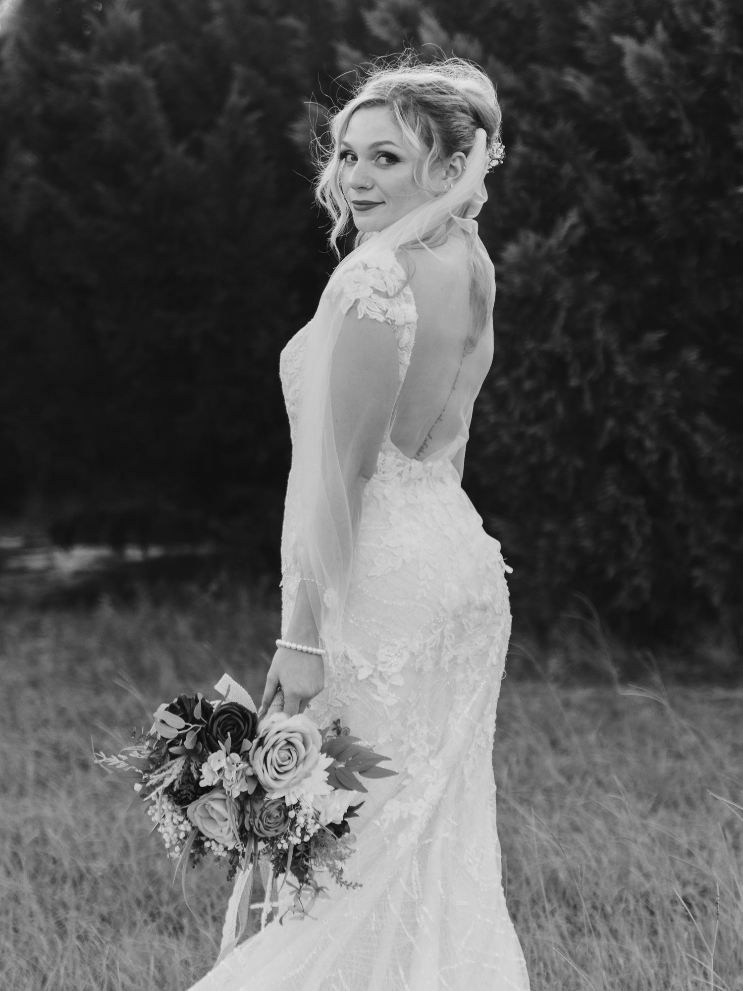 Black and white photo of bride in long-sleeve lace gown holding bouquet, looking over shoulder.