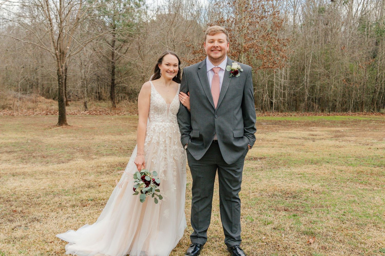 A couple in wedding attire poses together in a rustic outdoor setting with bare trees and grass in the background.