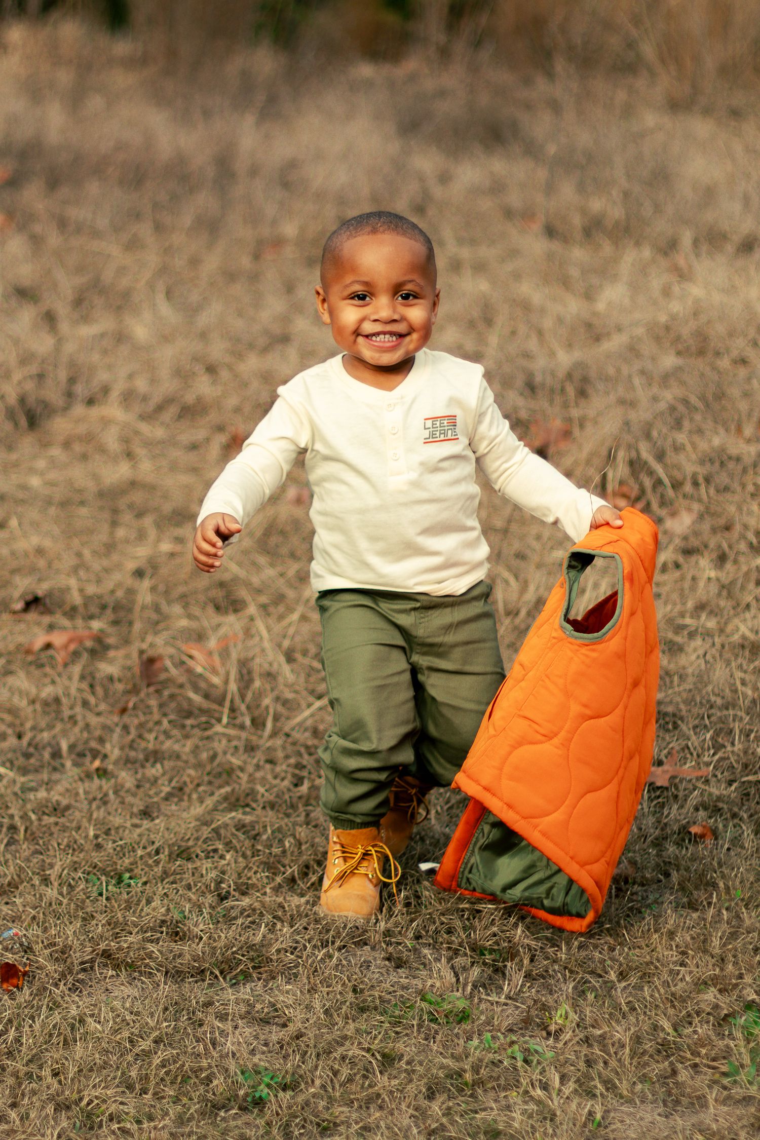 Small child in white shirt and green pants plays with orange pumpkin shaped bucket in autumn field.