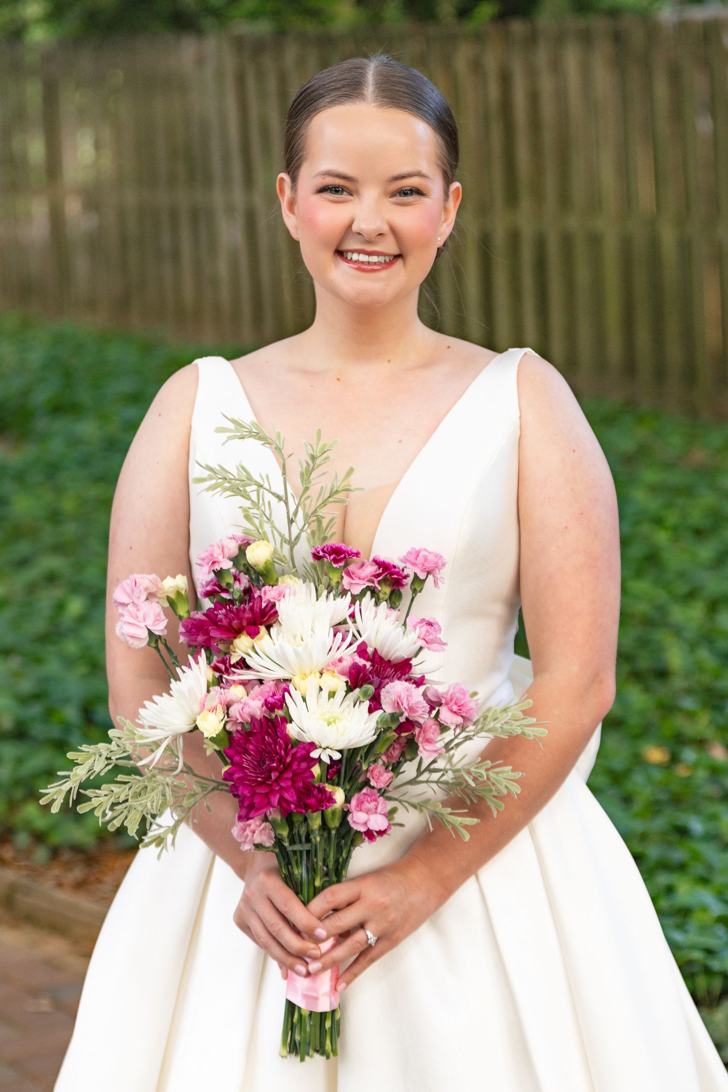 A bride in a white sleeveless wedding dress holds a vibrant bouquet of pink and white flowers in an outdoor garden setting.