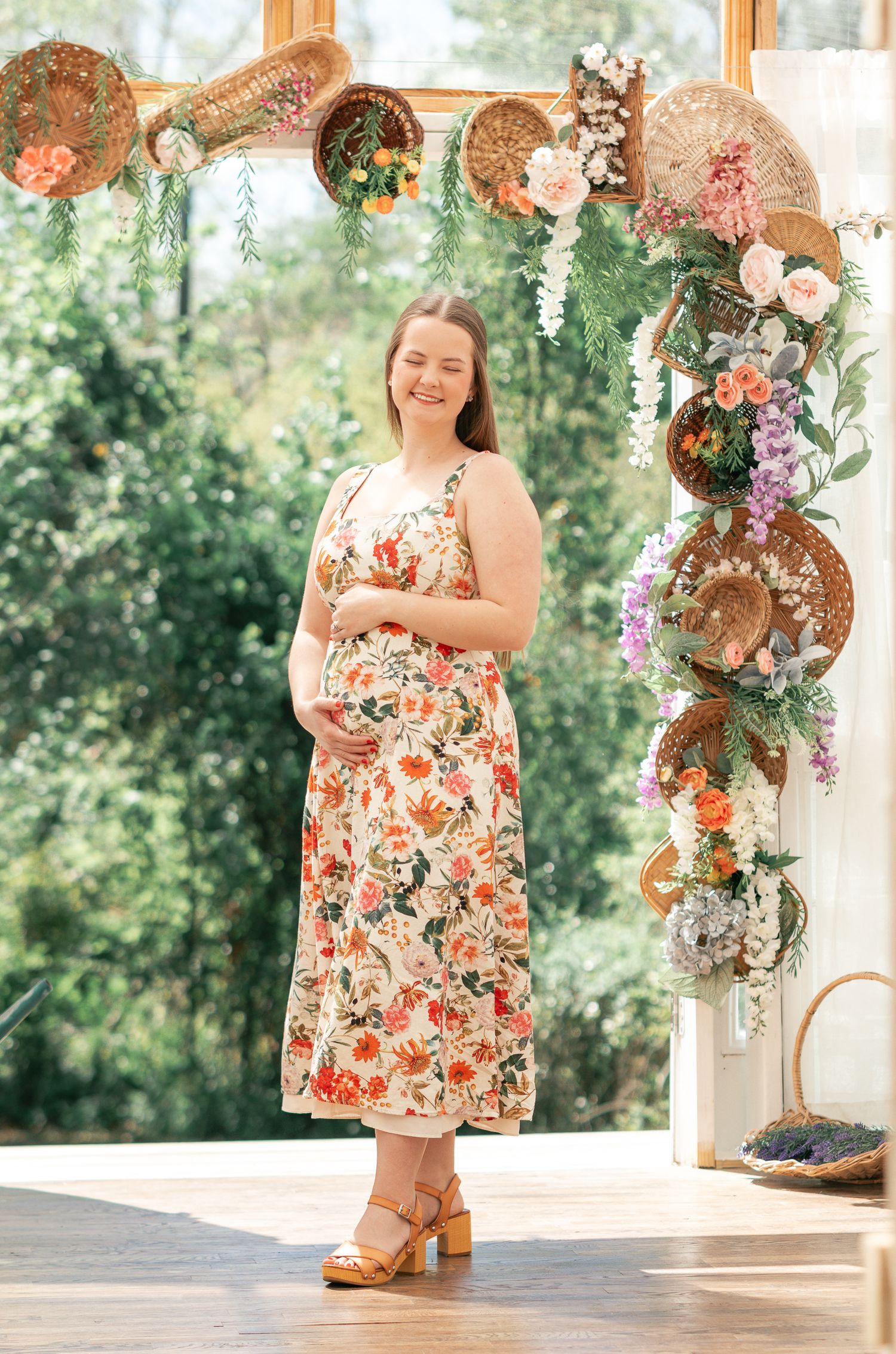 Pregnant woman in floral midi dress standing before decorative arch with wicker baskets and flowers.