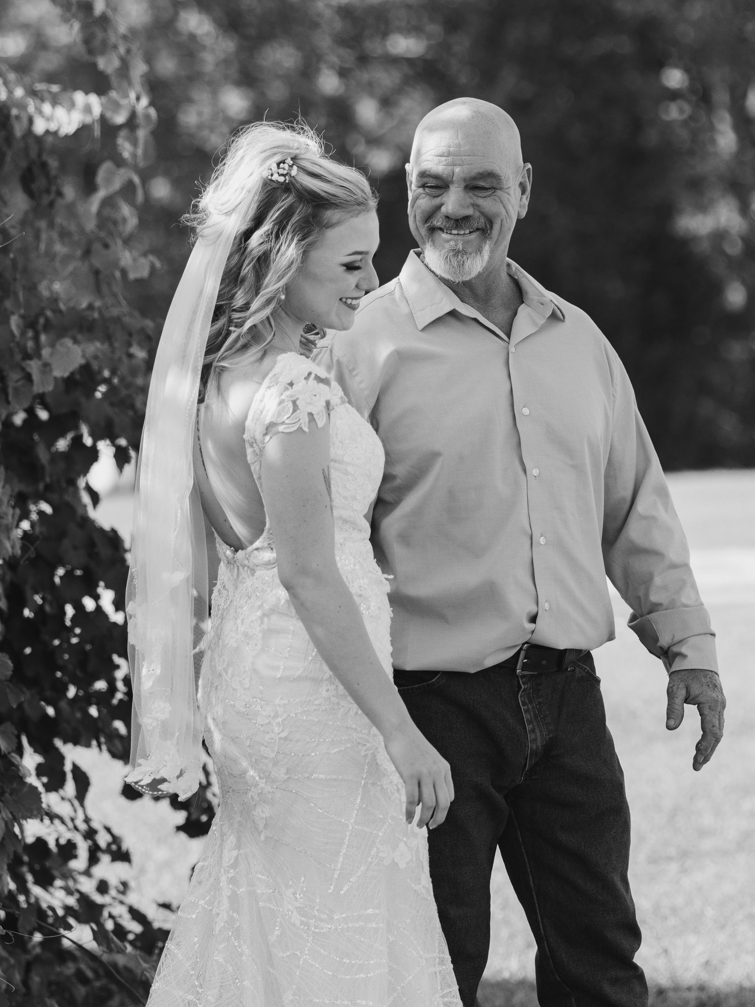 A smiling bride in a lace wedding gown and veil walks beside a bald man in a black and white outdoor photo.