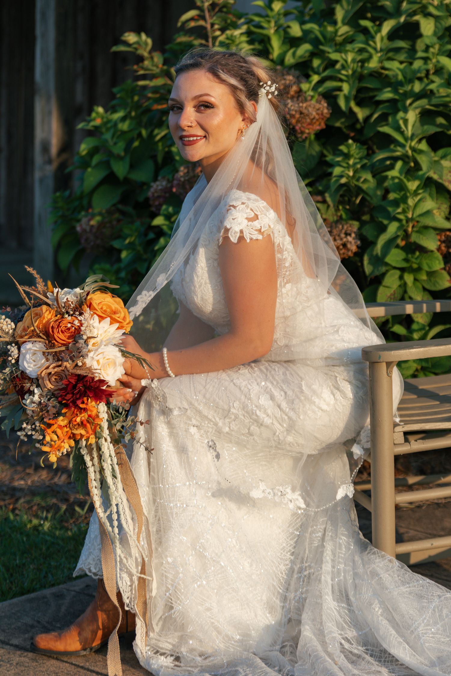 Bride in lace wedding dress sits on wooden bench holding vibrant orange and burgundy autumn bouquet at sunset.