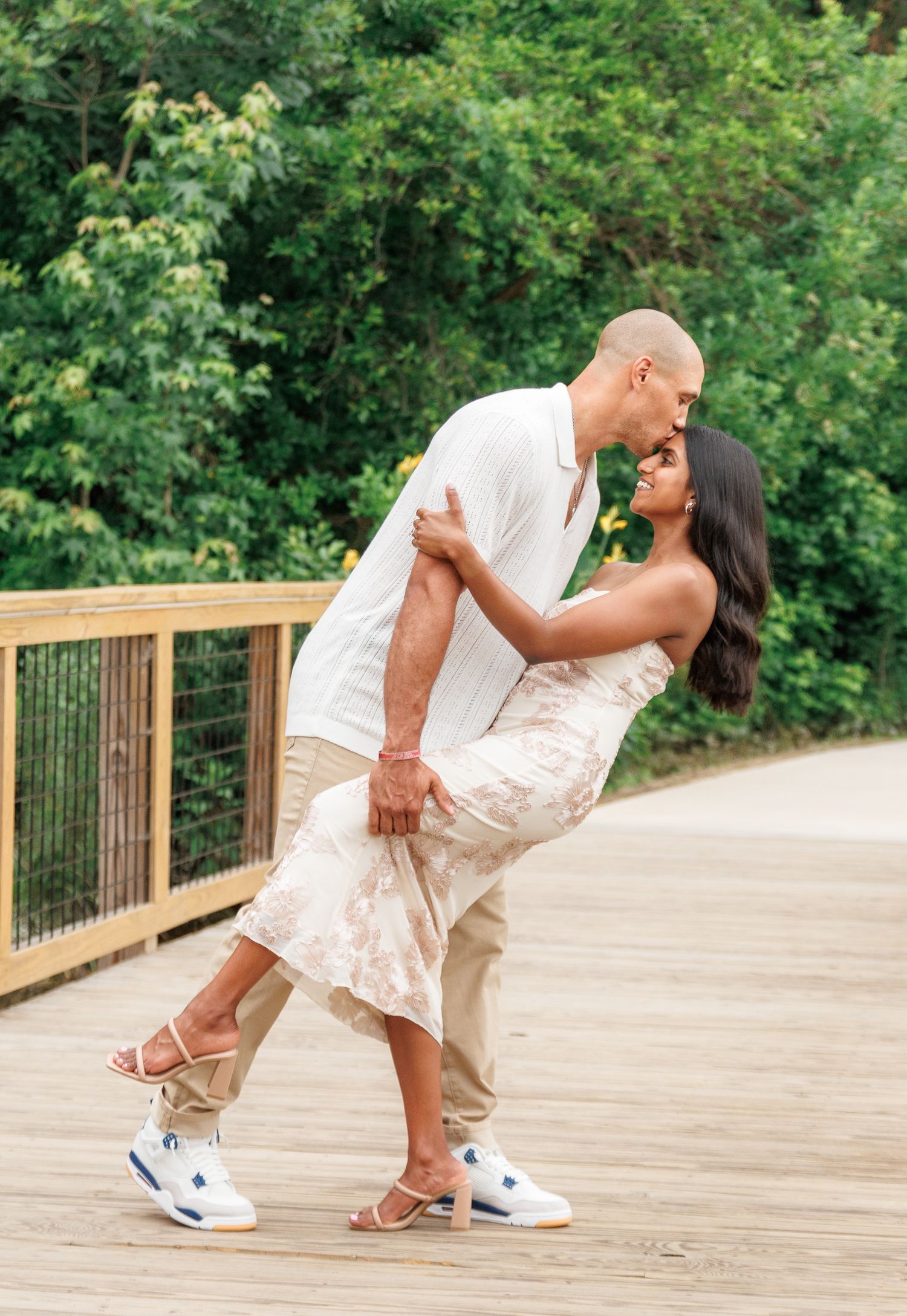 Couple sharing a romantic dip kiss on a wooden bridge surrounded by lush green trees.