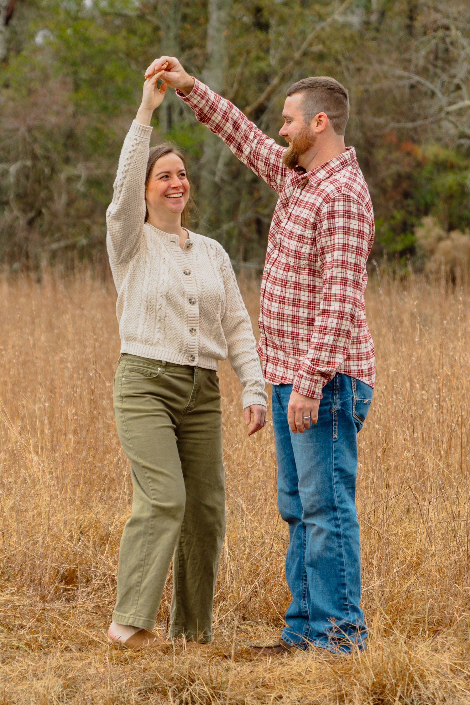 A man in a red plaid shirt twirls a smiling woman in a cream sweater in a golden autumn field.