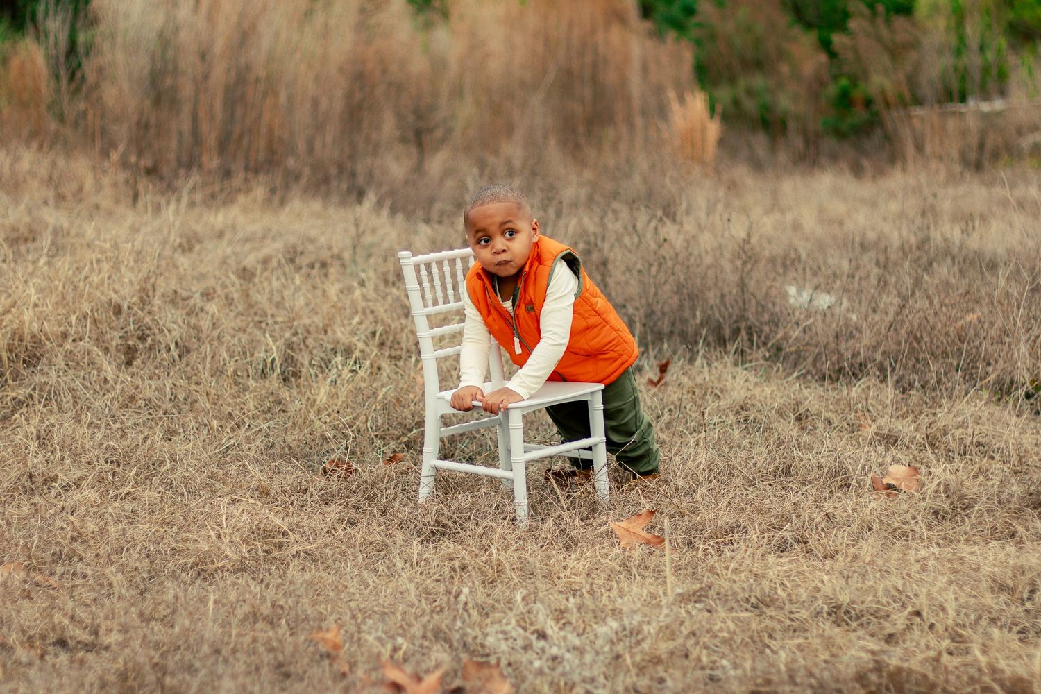 A white wooden chair sits in a brown autumn field with someone wearing an orange bandana and green pants.