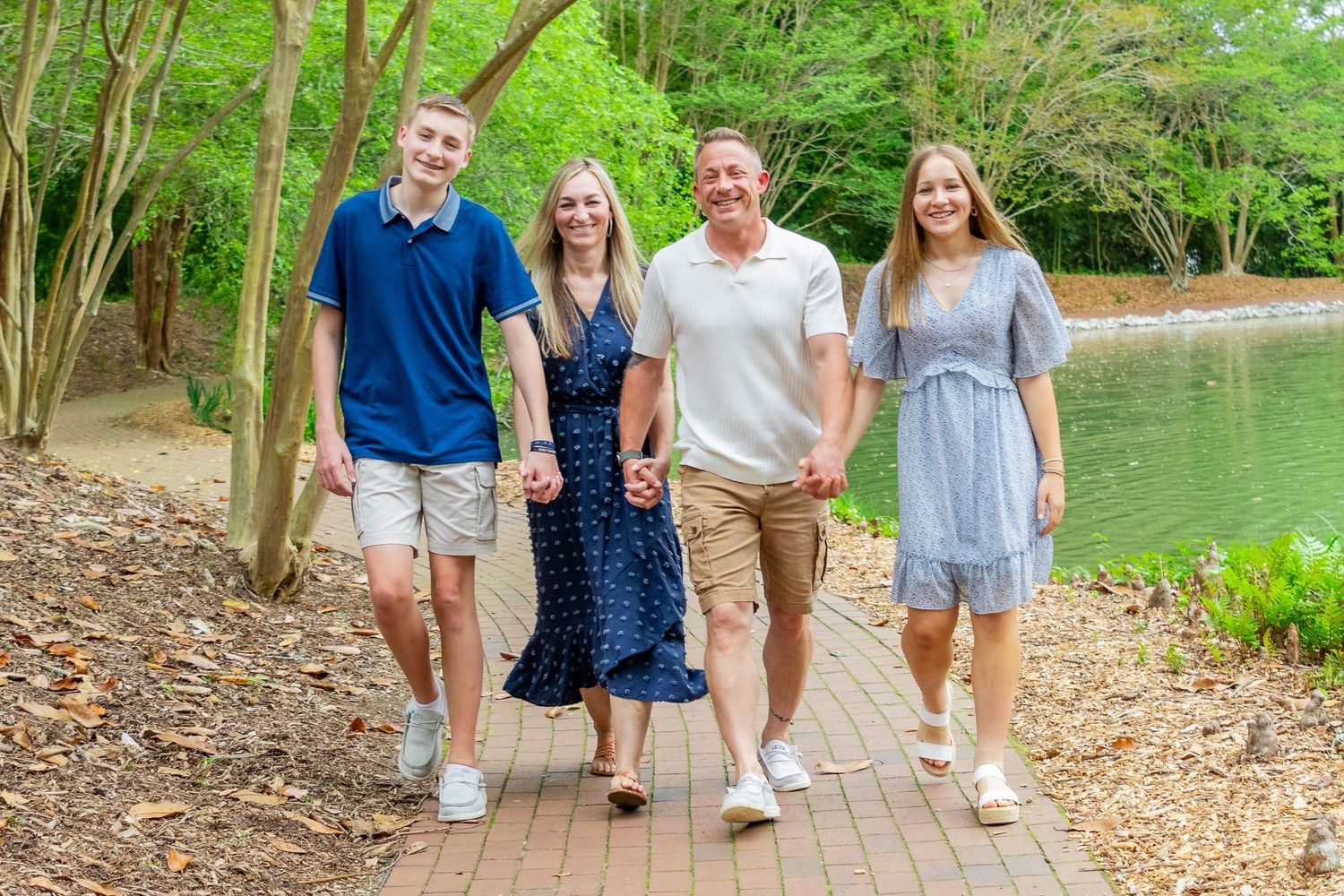 A family of four walks hand-in-hand along a scenic lakeside path surrounded by lush greenery.