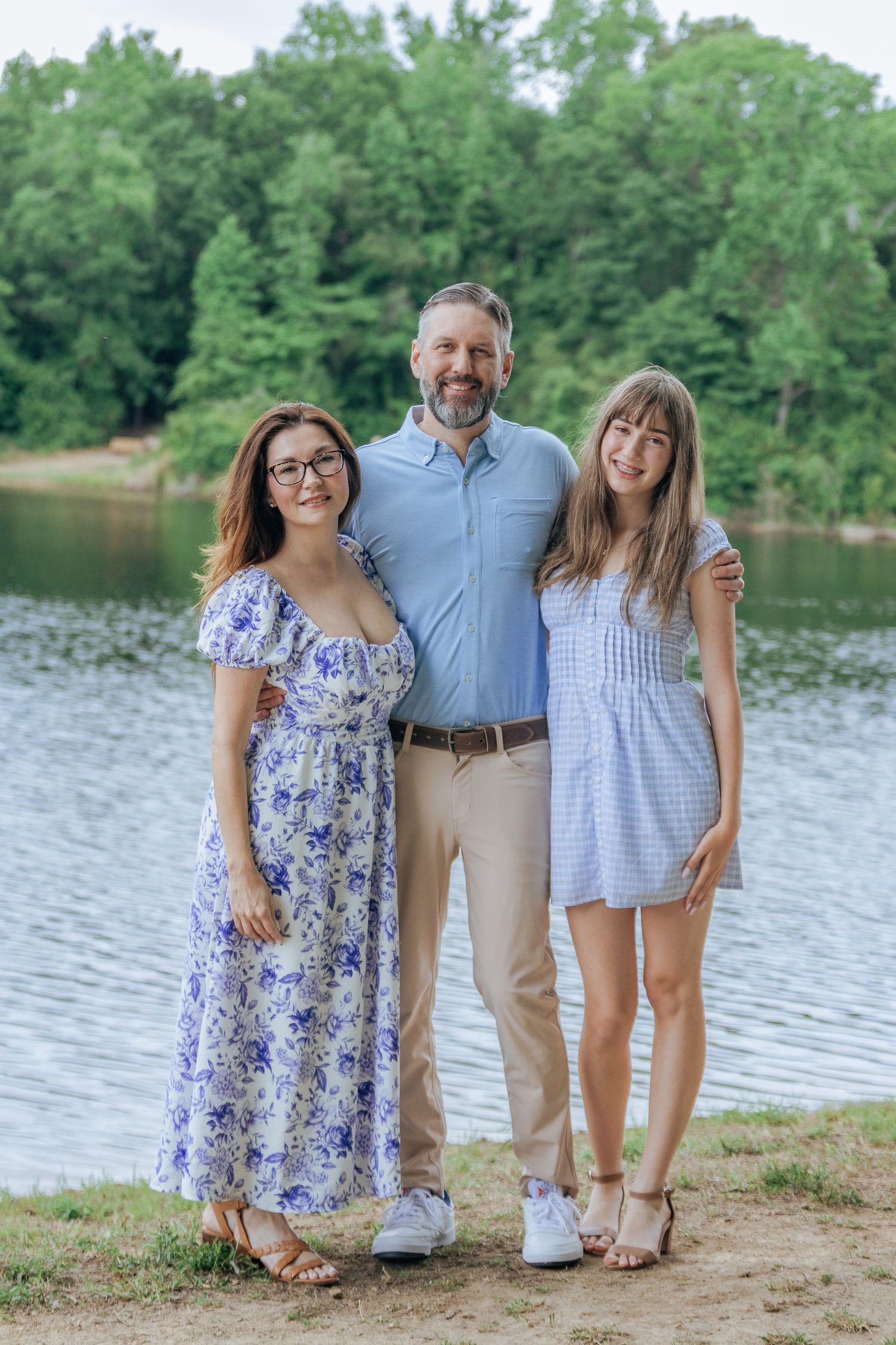 Family of three smiling together by a serene lake surrounded by green trees.
