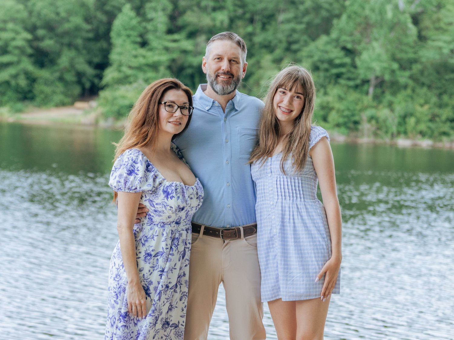 A man and two women smile together in front of a serene lake surrounded by lush green trees.