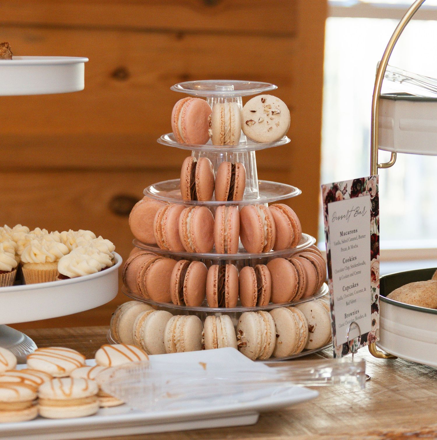 Pink and white macarons displayed on a tiered serving stand at a dessert table with wooden backdrop.