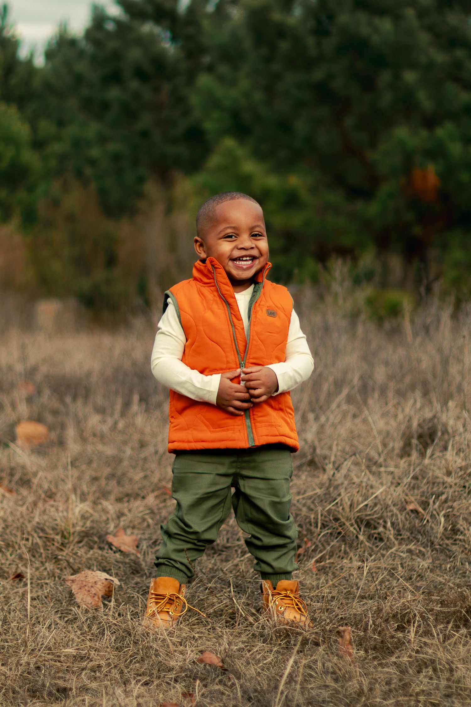 A young child wearing an orange vest and boots stands in a field of dried grass on an autumn day.