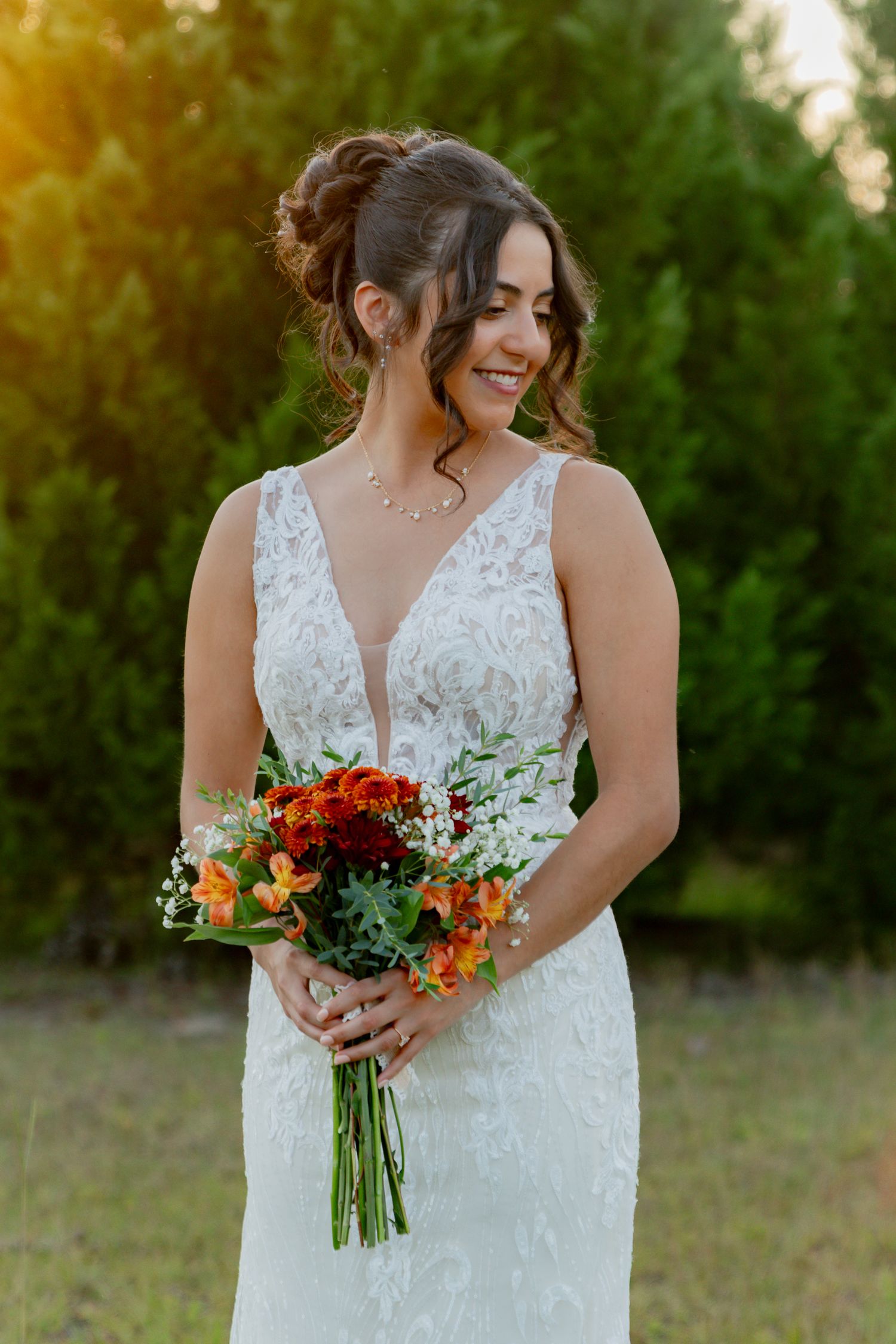 A person in a white lace wedding dress holds a vibrant red and orange floral bouquet in an outdoor setting.