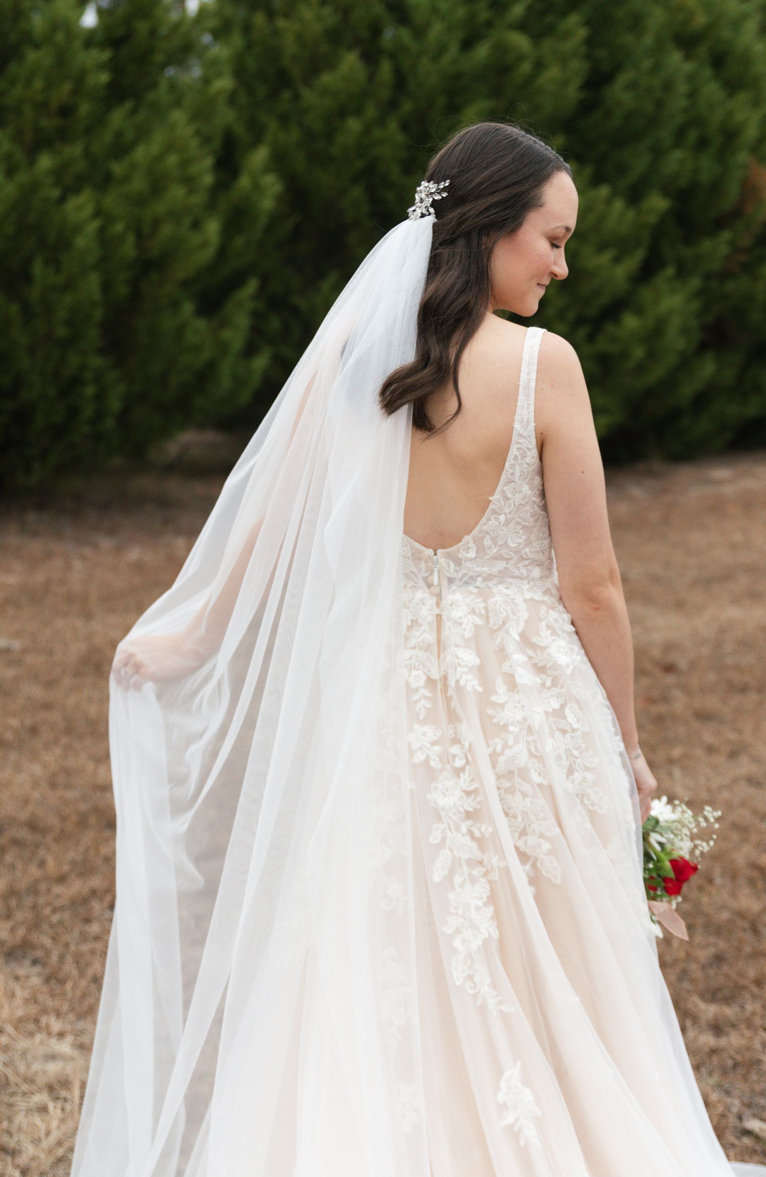 Bride in lace backless wedding gown with long cathedral veil standing near green trees.