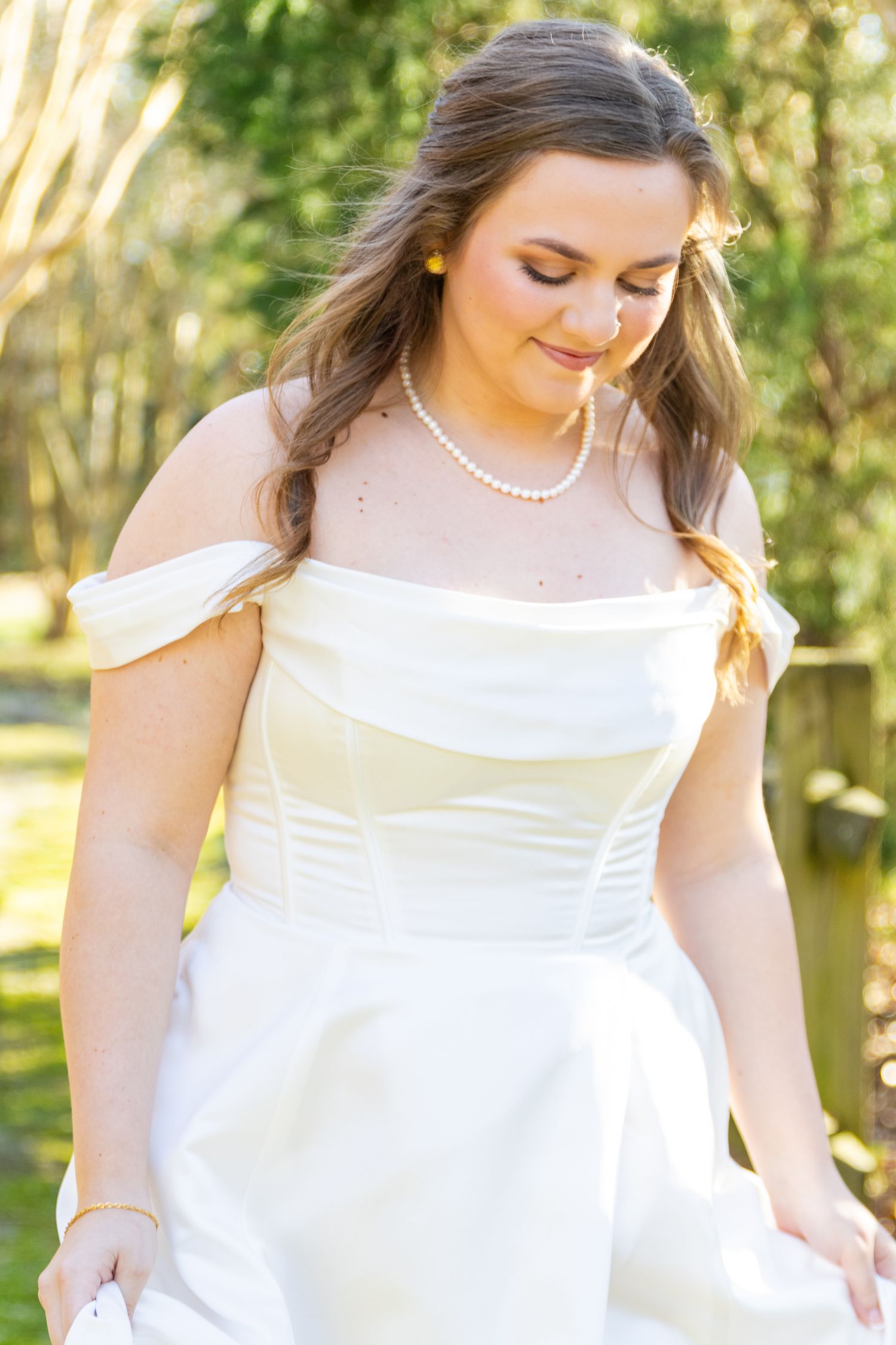Bride in off-shoulder ivory satin gown wearing pearl necklace looking down in sunny outdoor setting.