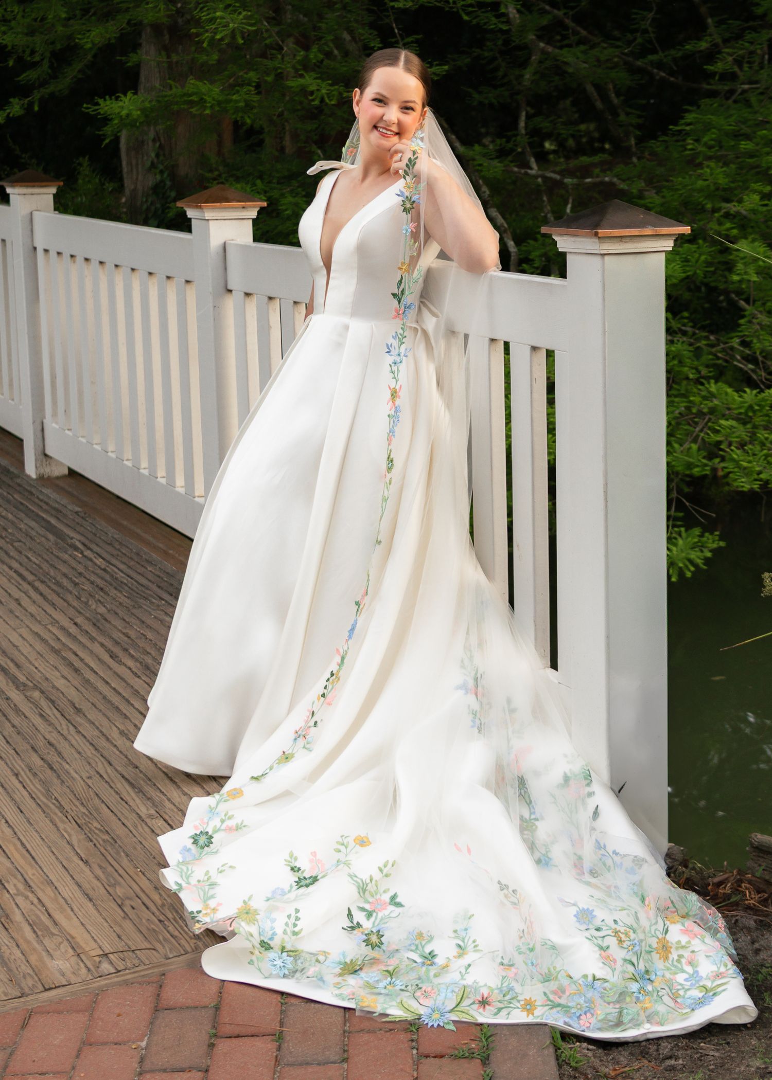White wedding dress with delicate floral embroidery along the hem displayed on a wooden deck with white railings.