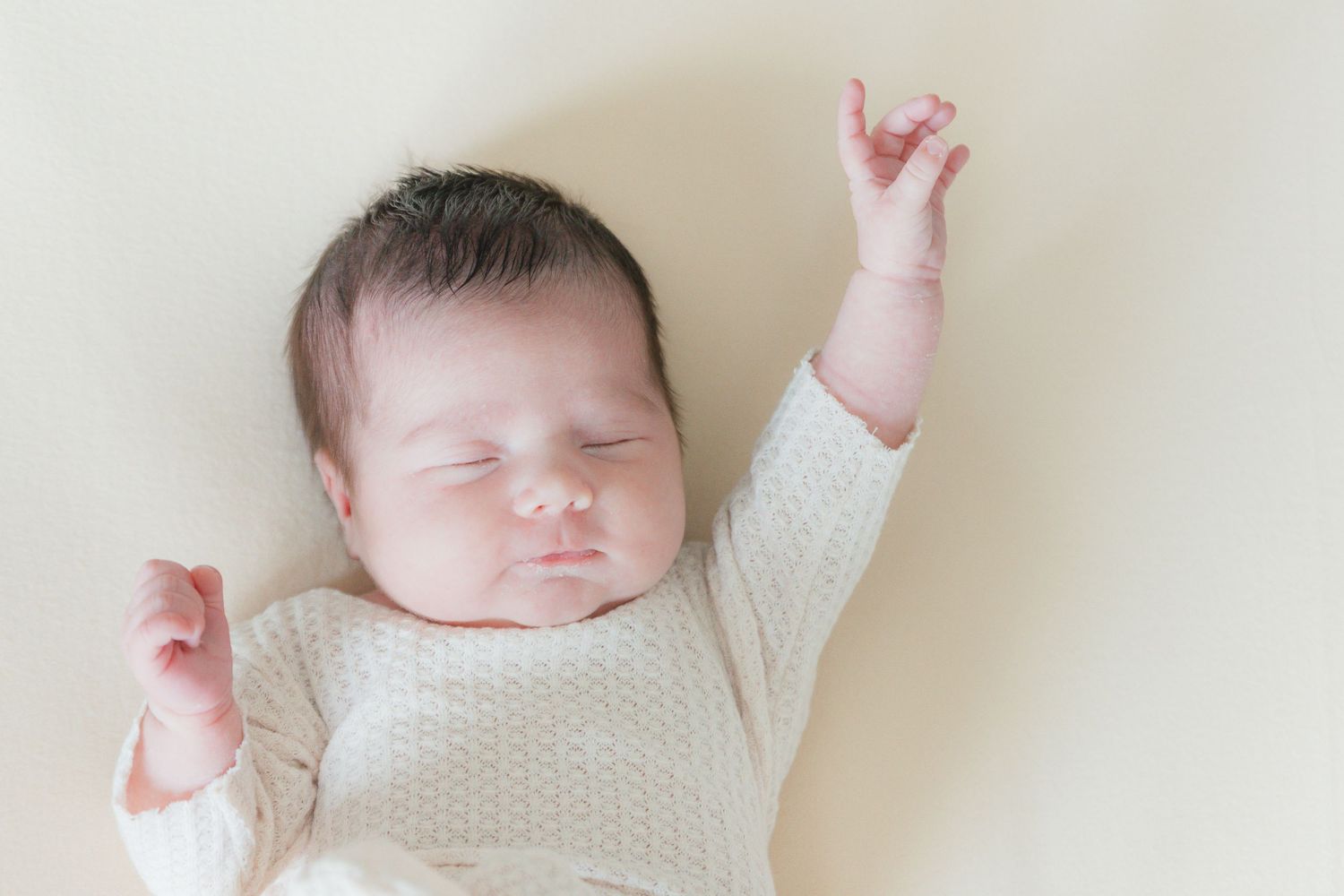 Peaceful newborn baby sleeping with arms raised, wearing a cream knit outfit on a soft beige background.