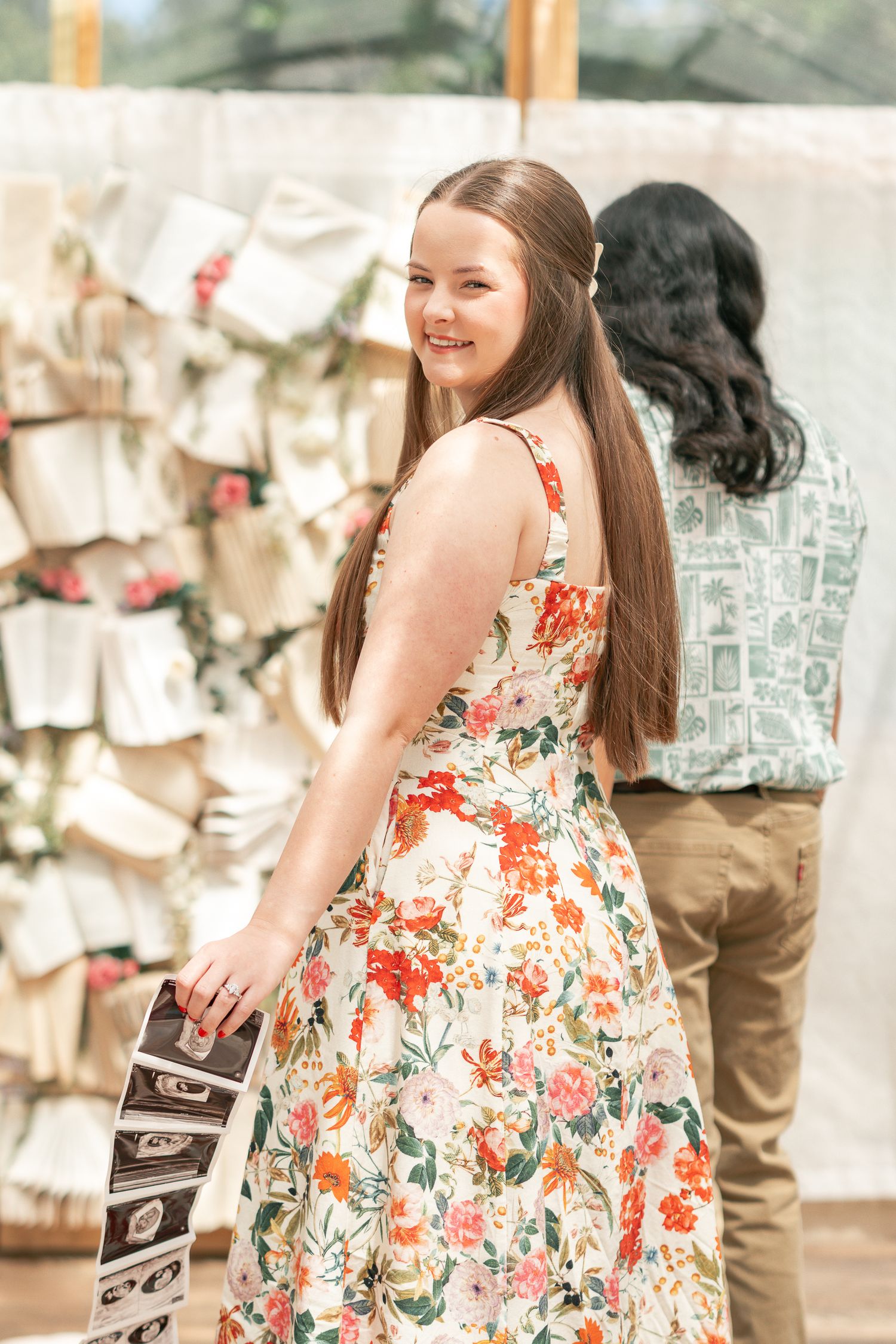 Young woman in floral sundress smiling over shoulder at outdoor wedding event with decorative card display.