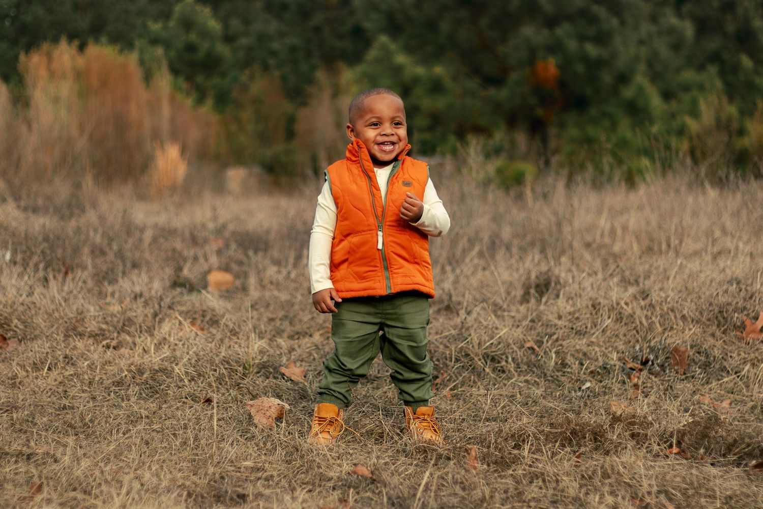 A child in an orange vest and green pants runs through a brown grassy field on a sunny day.