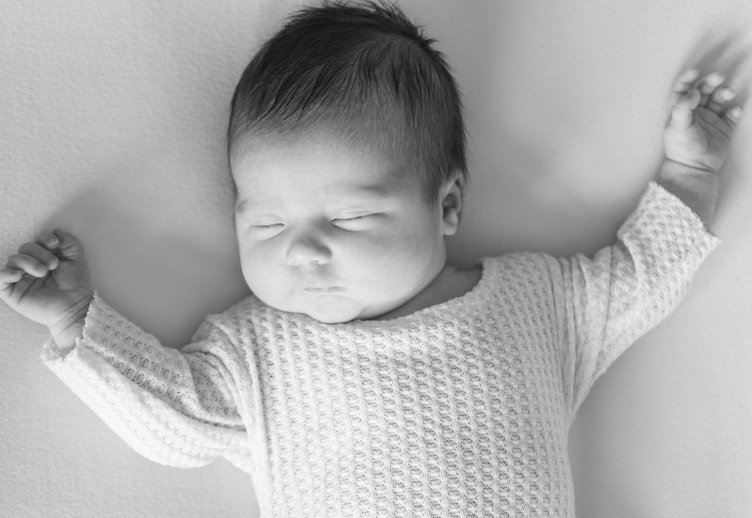 Black and white photo of a sleeping newborn baby in a knit onesie with arms stretched out.